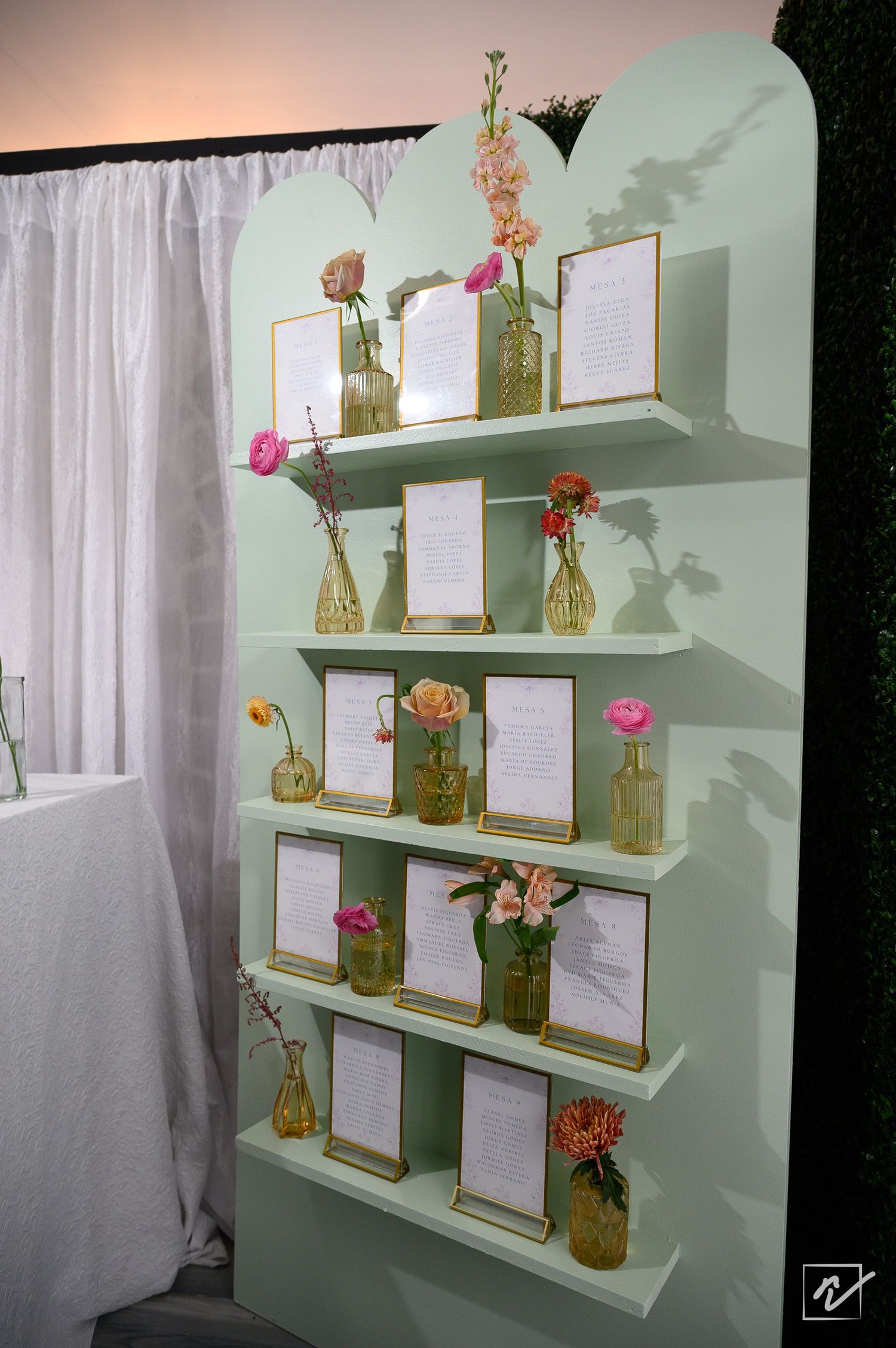 Mint green wedding seating chart display with tiered shelves holding framed table cards and small gold vases of pink and coral flowers.