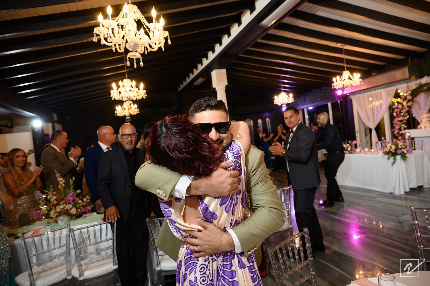 Groom in sunglasses and a green suit hugs a woman in a purple dress at a wedding reception under chandeliers.