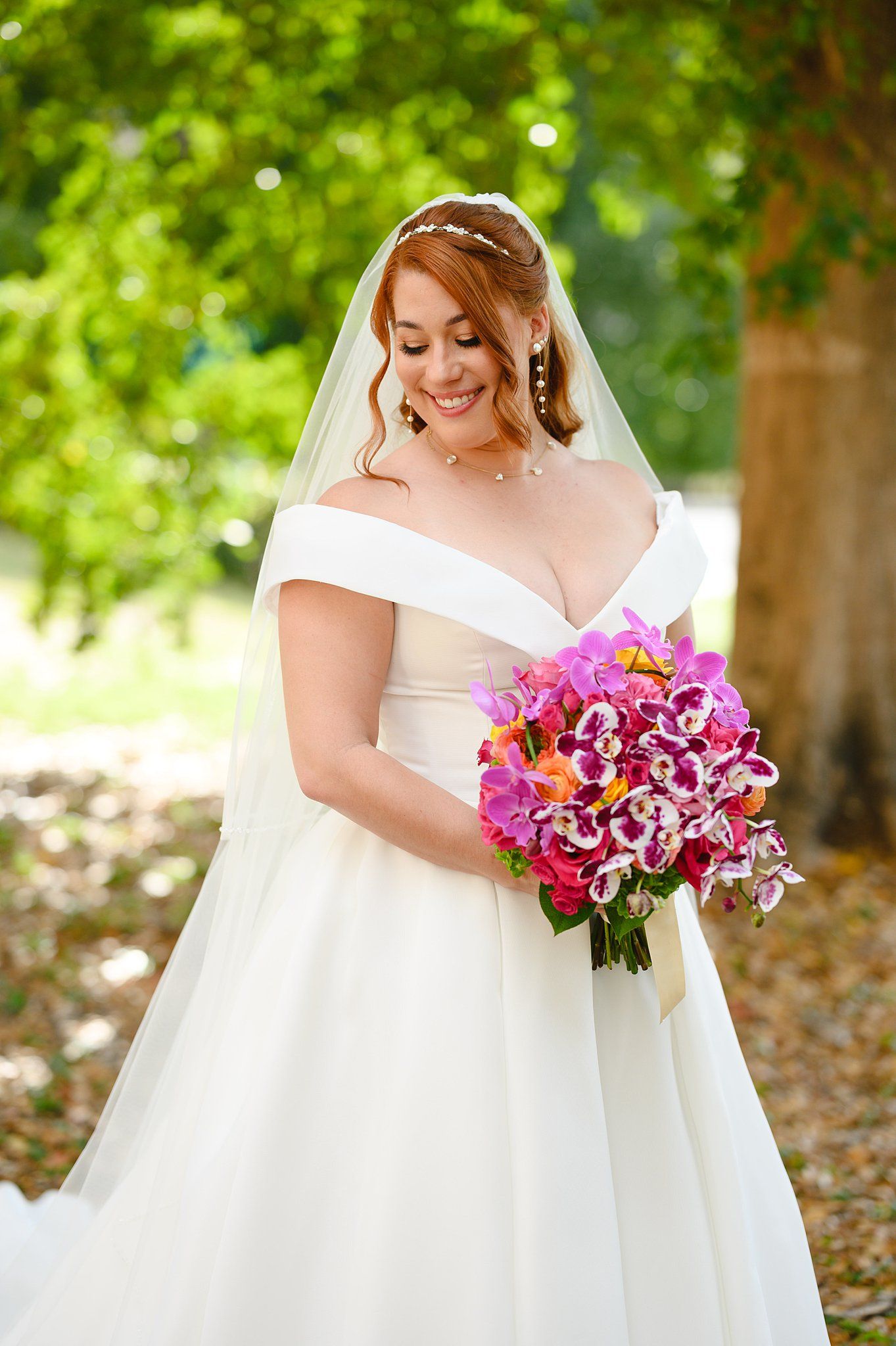 Smiling bride in off-shoulder white gown and veil holds vibrant pink orchid bouquet outdoors among lush green trees.