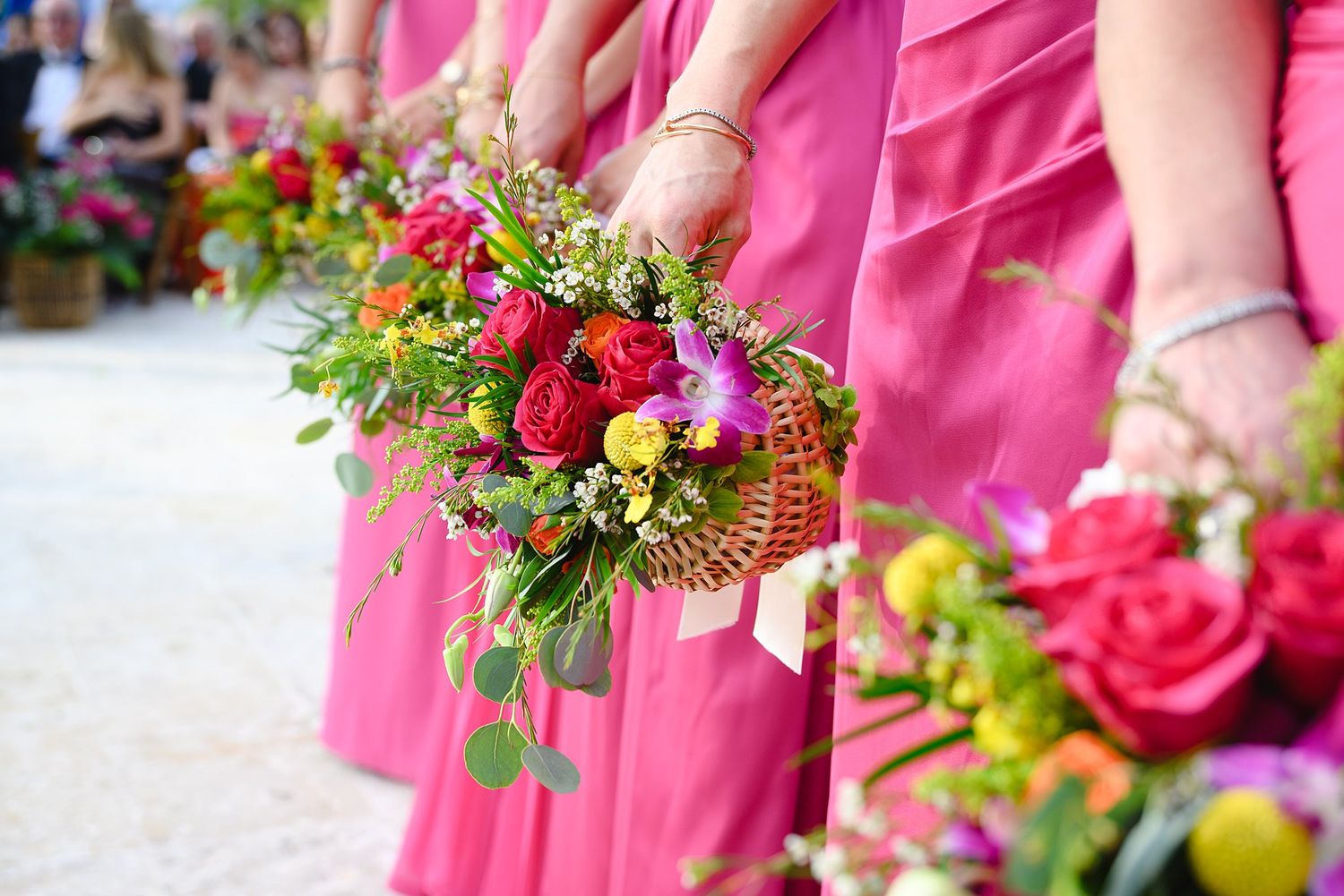 Bridesmaids in pink dresses holding colorful floral bouquets in wicker baskets at an outdoor wedding ceremony.