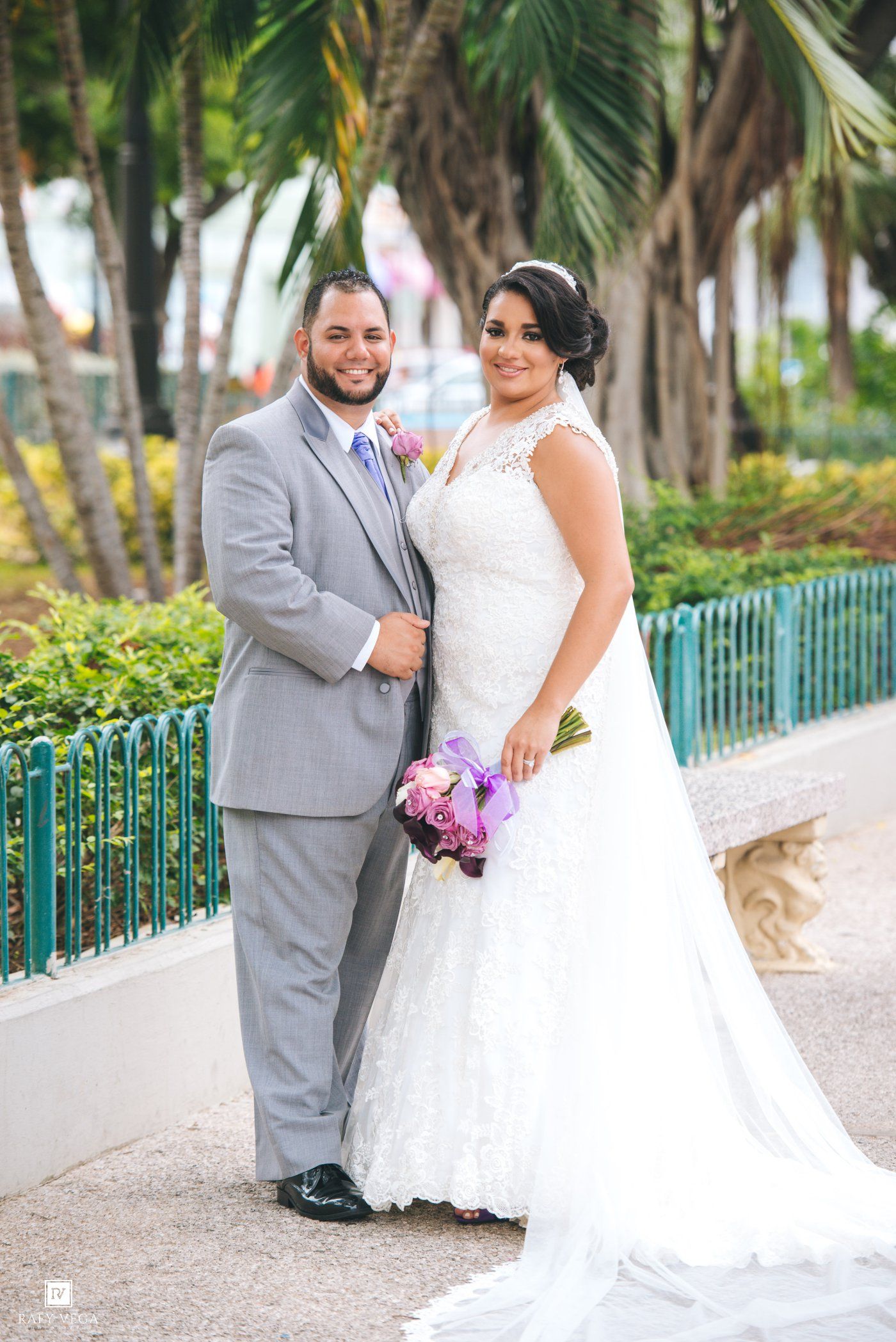 Bride in white lace gown holding purple bouquet stands with groom in gray suit outdoors at tropical wedding venue.