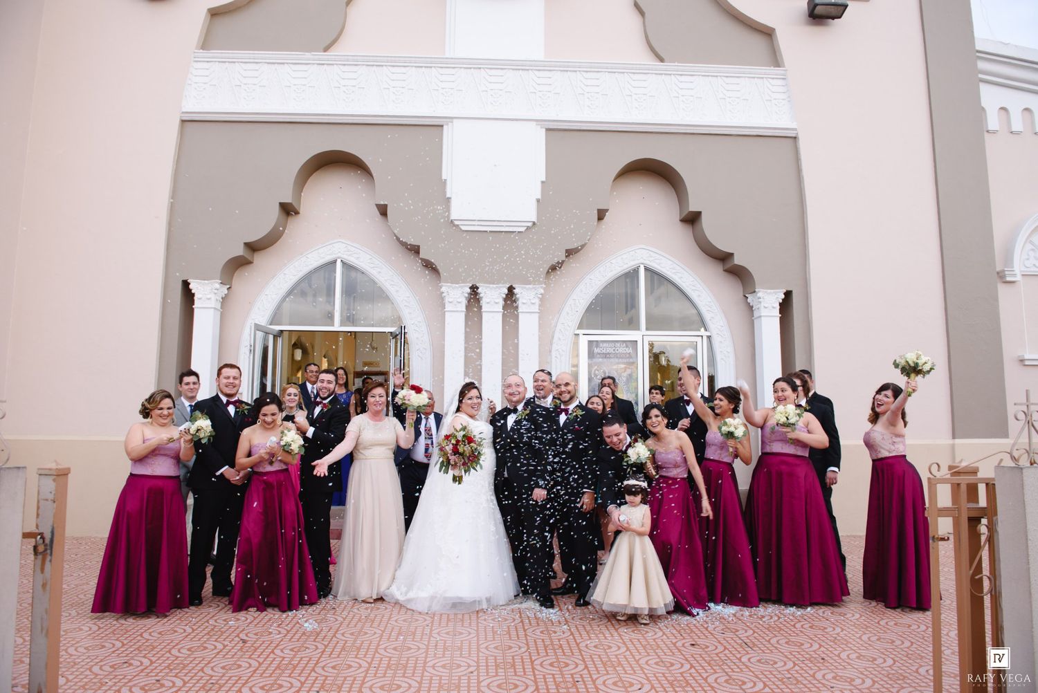 Large wedding party in burgundy and champagne attire poses outside an elegant venue with arched windows.