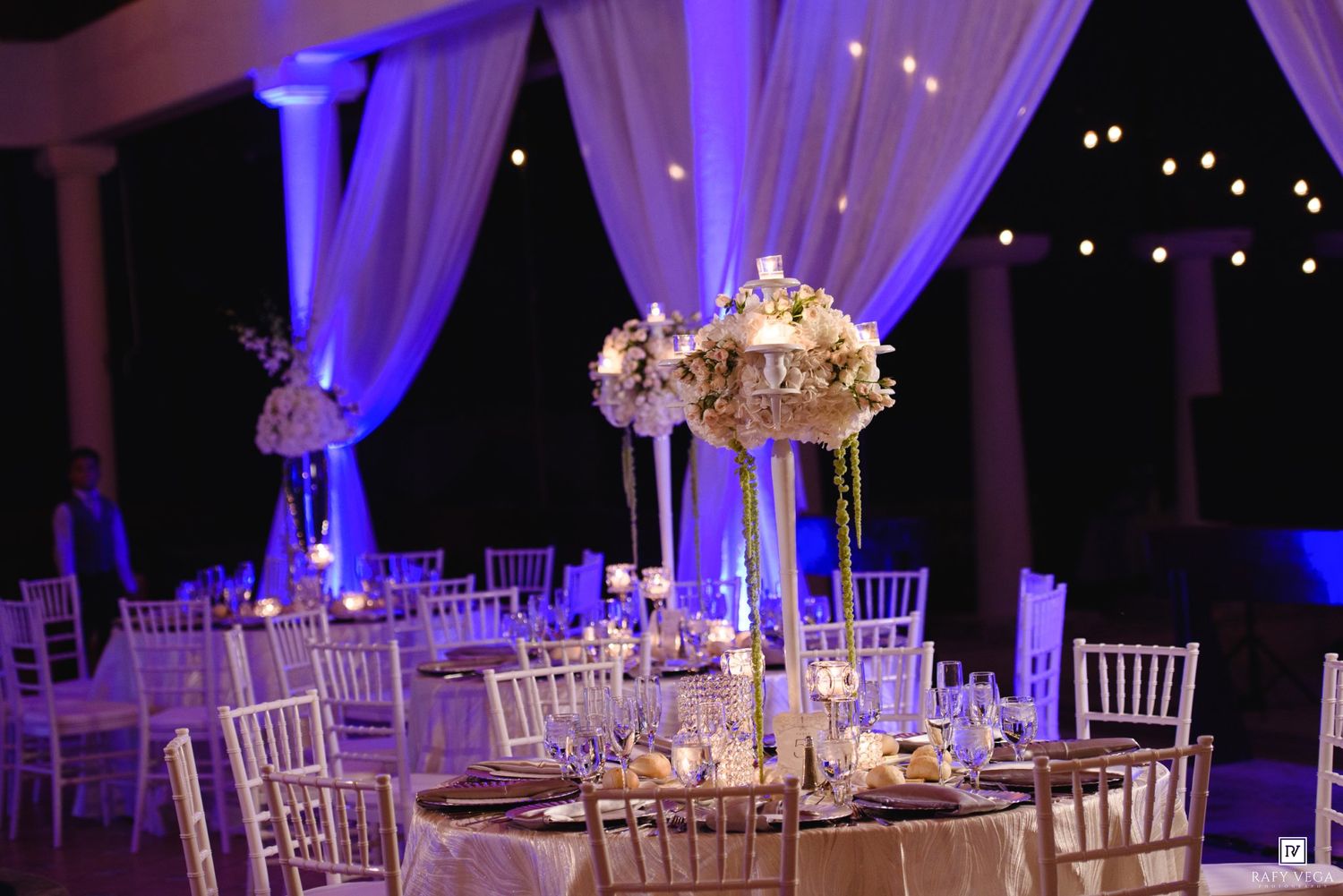 Elegant wedding reception with tall floral centerpieces, white chiavari chairs, candlelit tables, and purple uplighting.