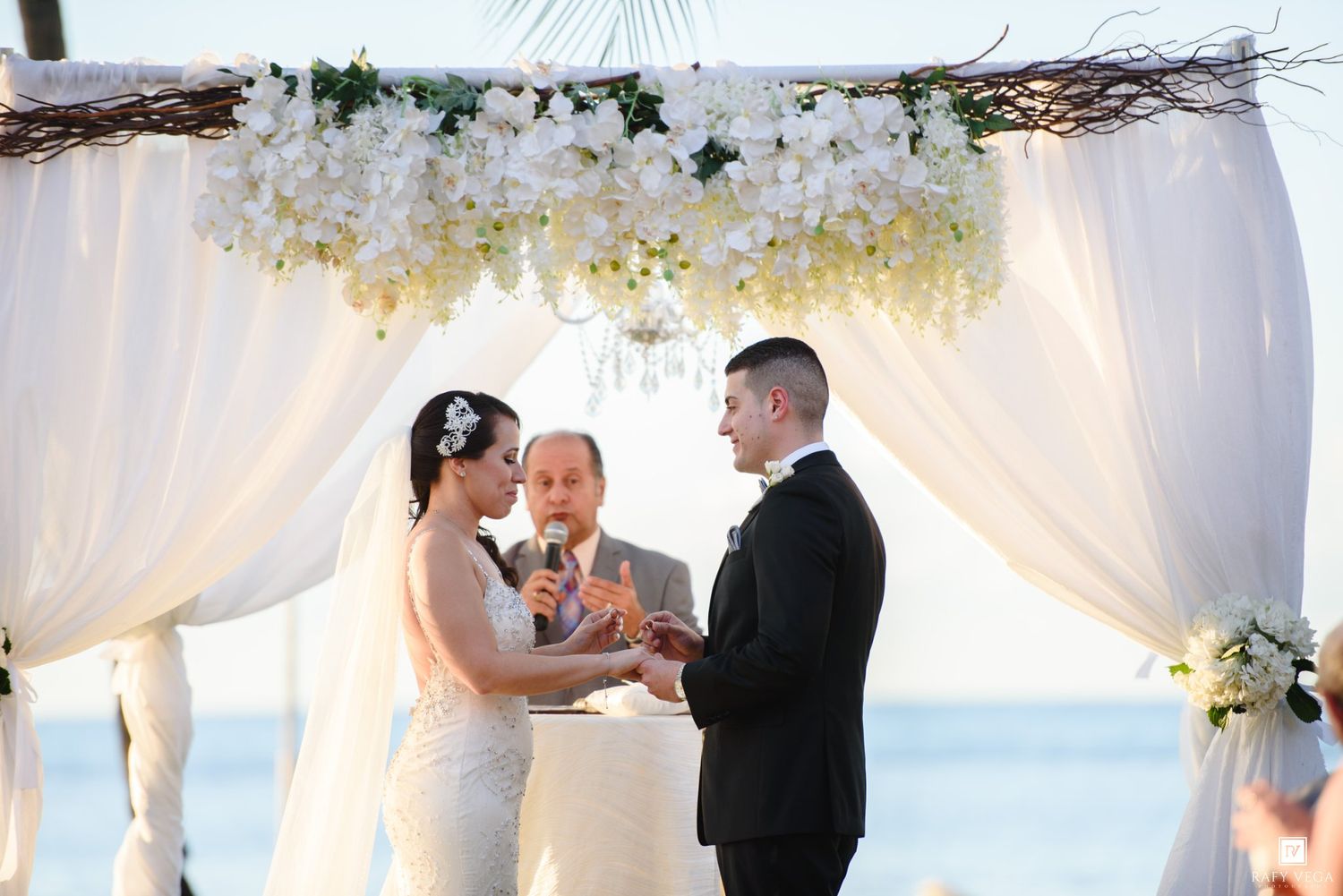 Couple exchanging vows at a beach wedding ceremony under a white floral arch with ocean backdrop.