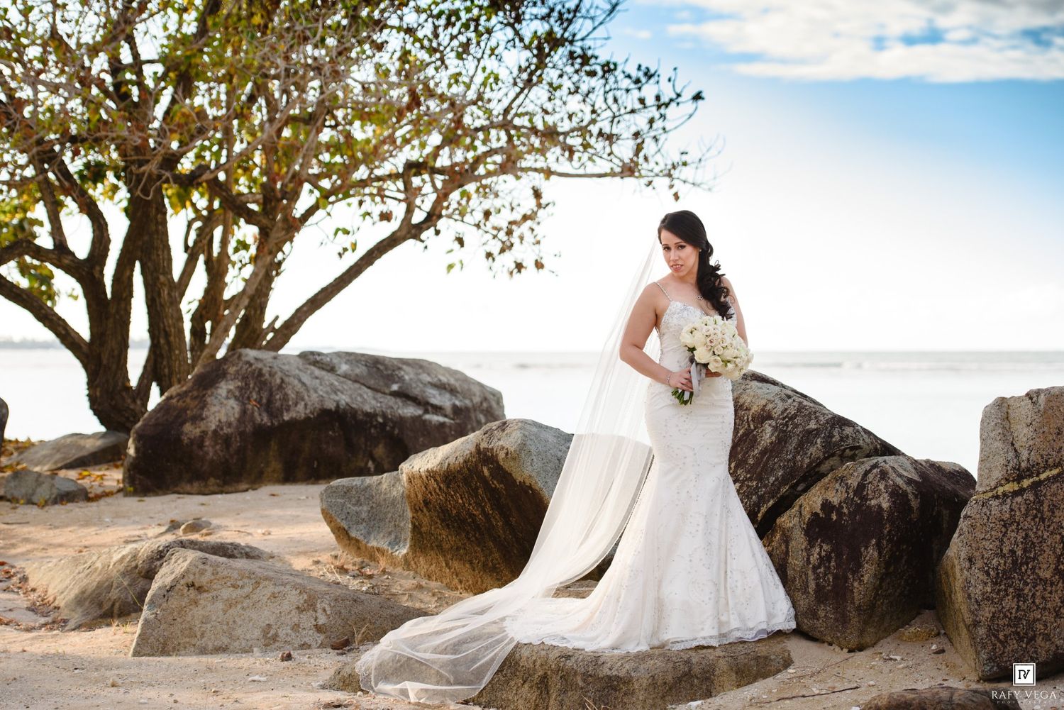Elegant bride in lace mermaid dress holds bouquet beside large rocks on a scenic tropical beach.