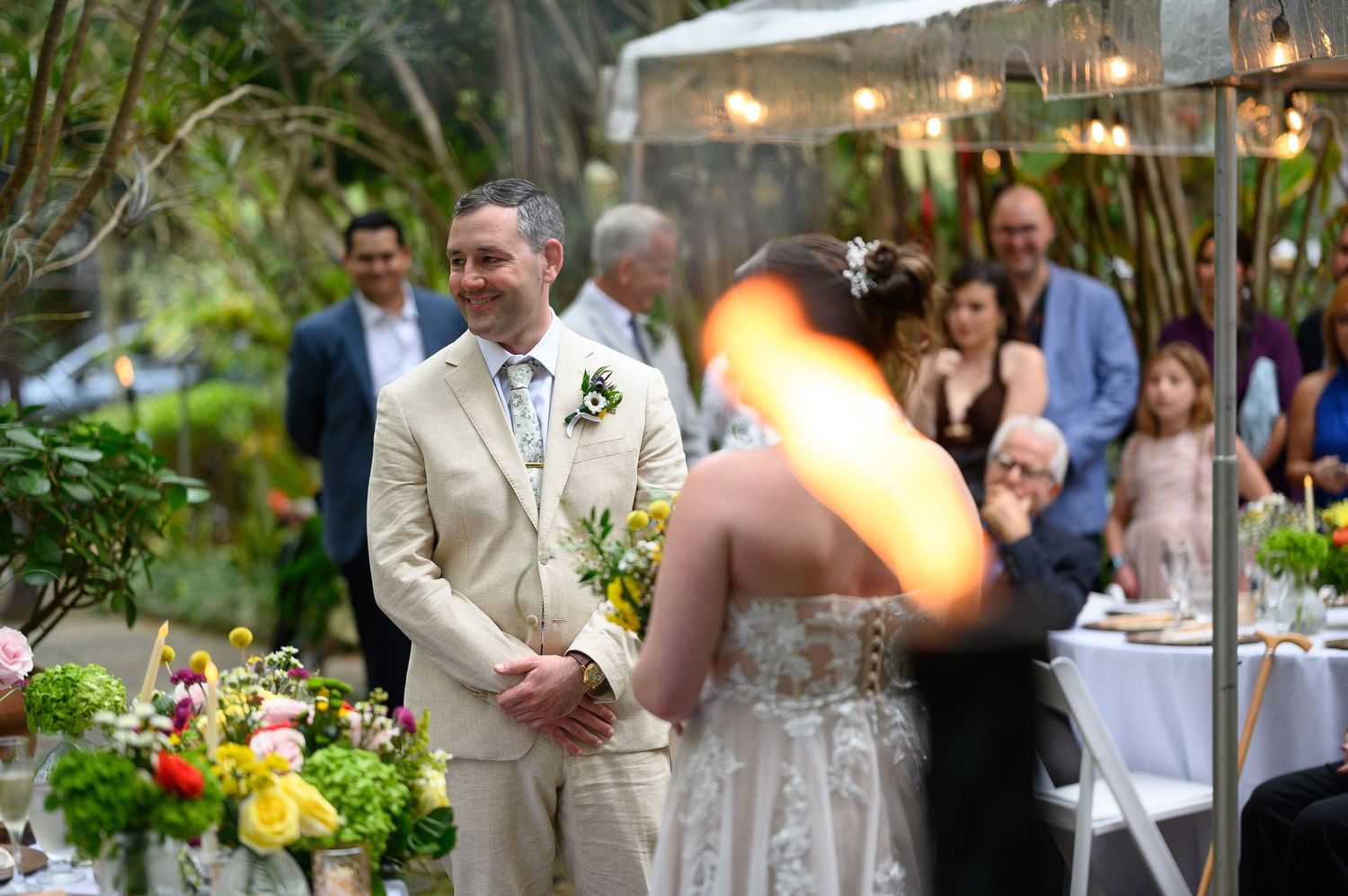 Groom in beige suit smiles at bride during outdoor garden wedding ceremony with floral arrangements and string lights.