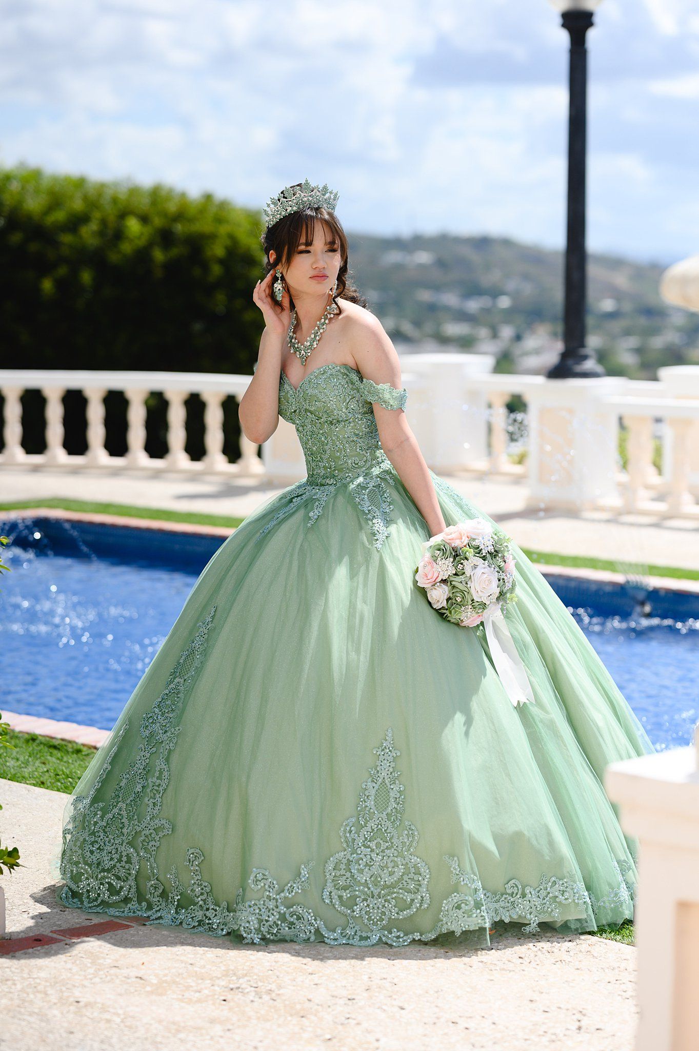 Young woman in sage green quinceañera ball gown with lace details, tiara, and pink bouquet poses by a pool.