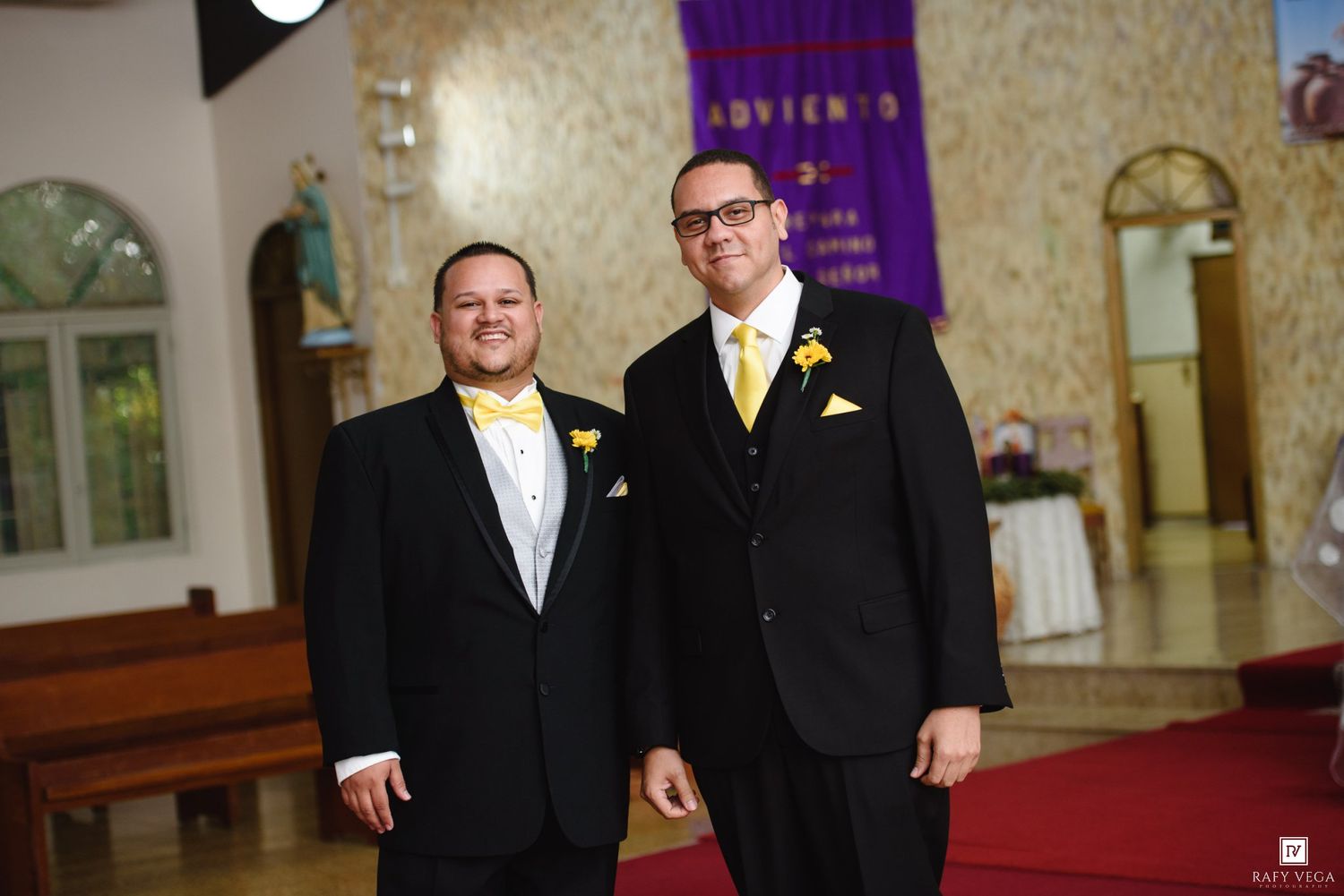 Two men in black suits with yellow bow ties and boutonnieres smile together inside a church before a wedding ceremony.