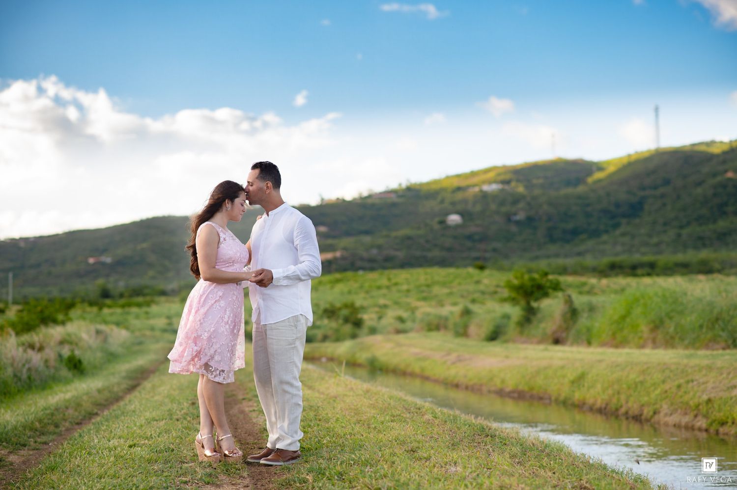 Couple in pink and white attire sharing a romantic moment on a lush green field with scenic hills backdrop.