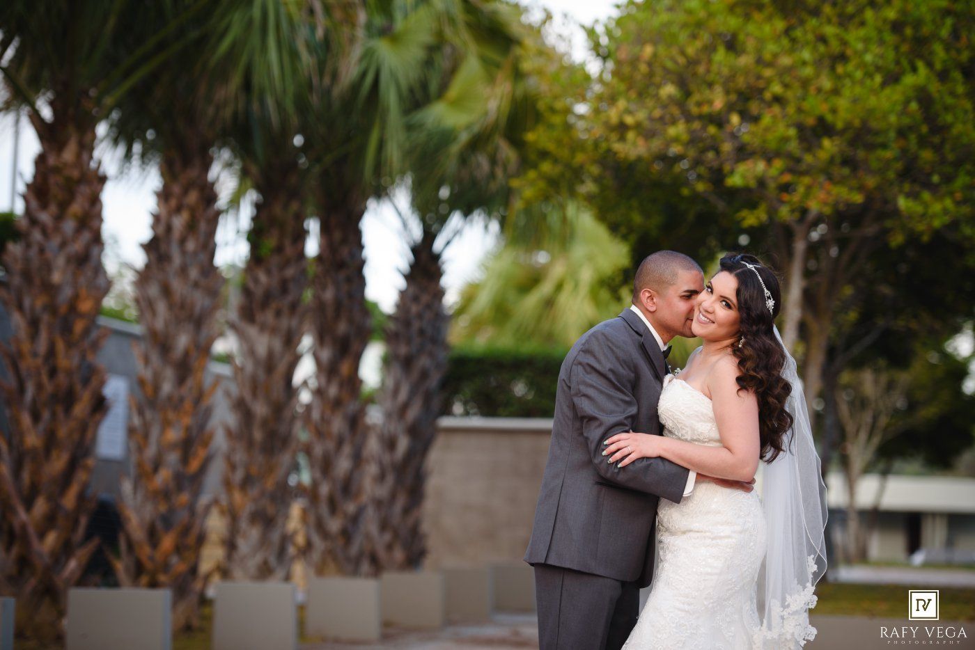 Bride and groom sharing a romantic kiss outdoors with palm trees in the background on their wedding day.