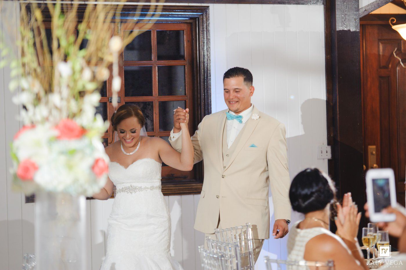 Newlyweds make a grand entrance at their wedding reception, groom in tan suit with teal bow tie, bride in lace gown.