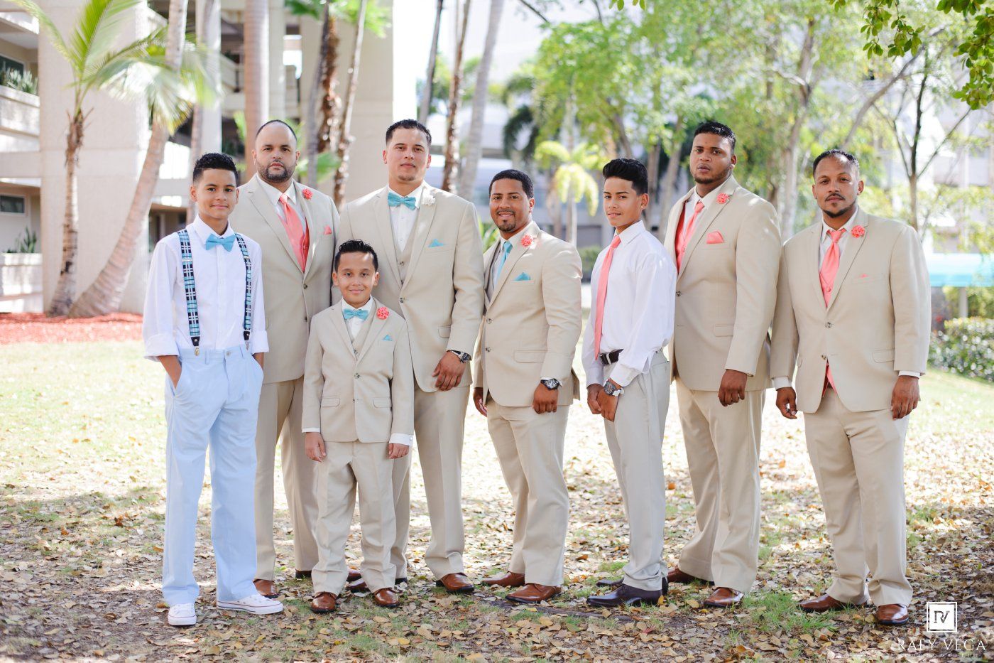 Groomsmen in tan suits with coral ties pose outdoors at a tropical wedding venue with lush greenery.