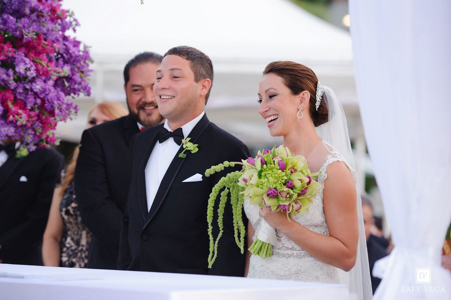 Smiling bride holding green and pink bouquet stands with groom in tuxedo at elegant outdoor wedding ceremony.