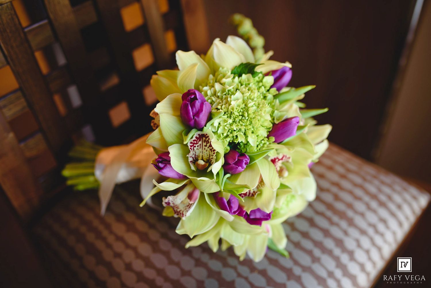 A bridal bouquet featuring green cymbidium orchids, purple tulips, and lime green blooms on a patterned chair.