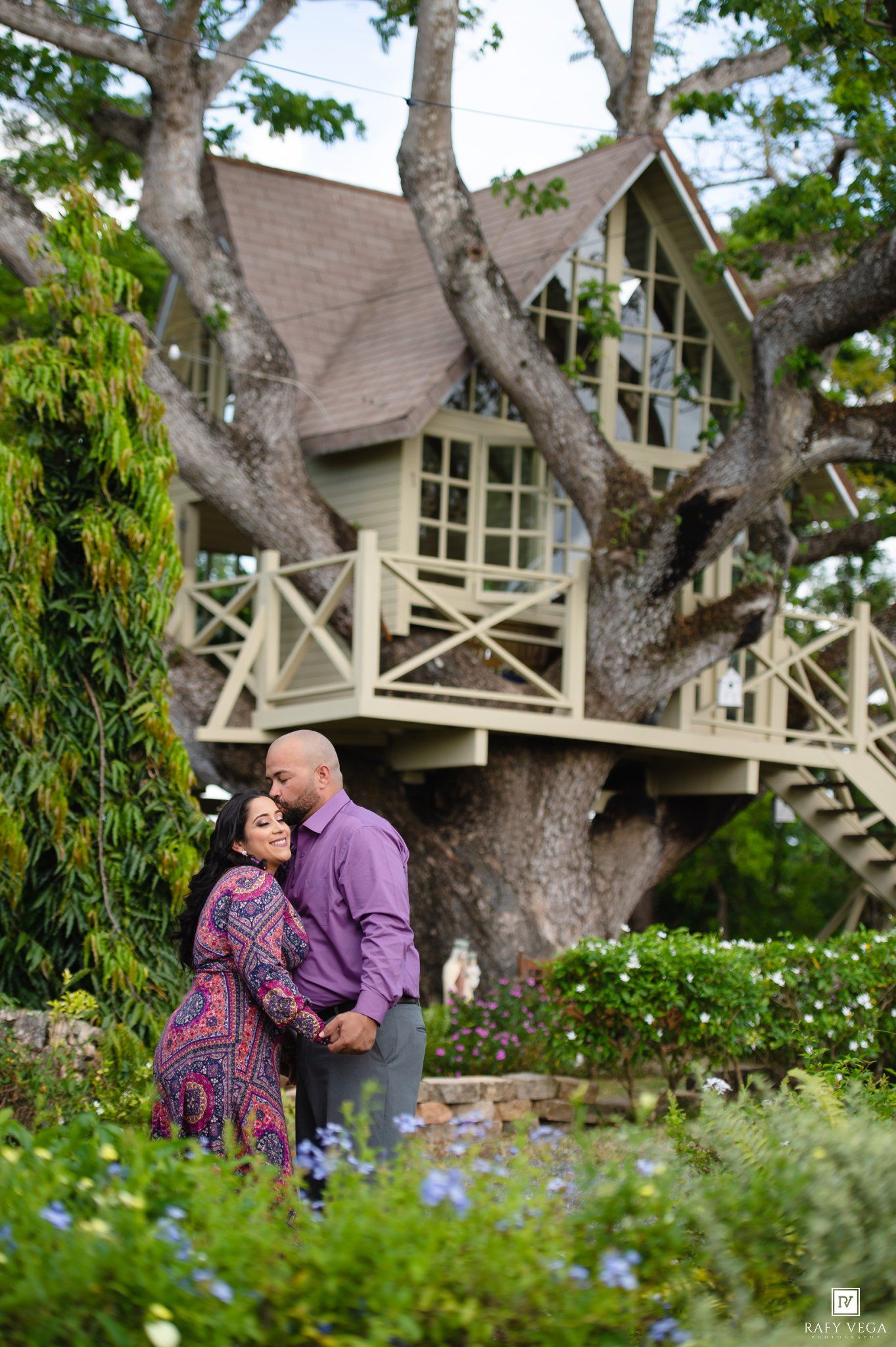 Couple embracing romantically in a lush garden with a charming treehouse backdrop during an outdoor engagement photo session.