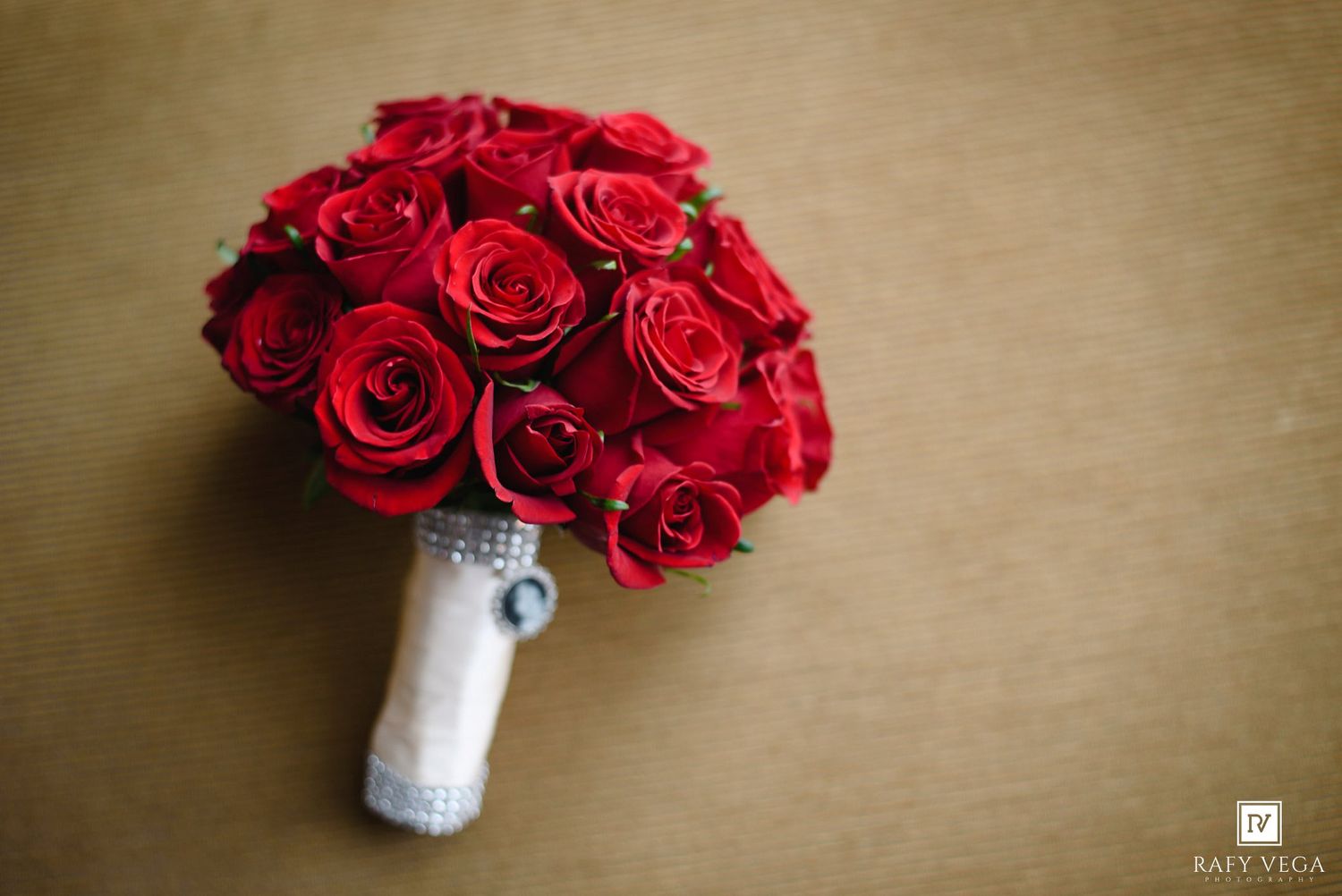 A stunning bridal bouquet of vibrant red roses with a silver ribbon handle on a golden background.