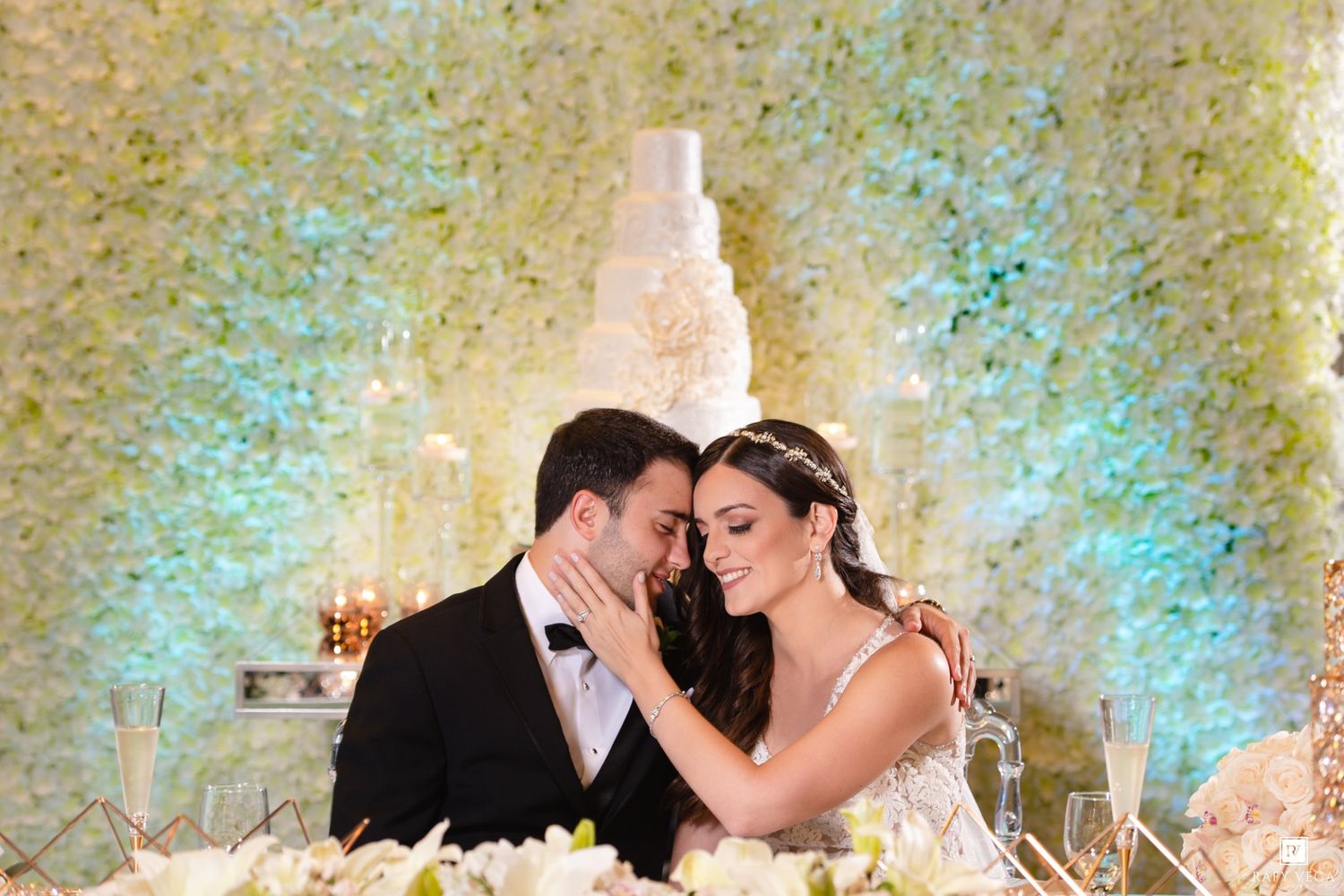 Bride and groom share a romantic moment at wedding reception with white floral backdrop and elegant tiered cake.