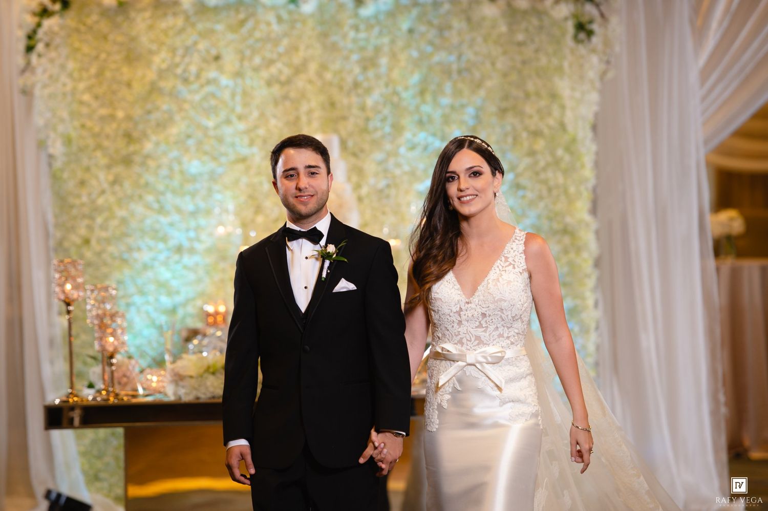 Bride and groom holding hands at wedding altar with floral backdrop, bride in white gown, groom in black tuxedo.