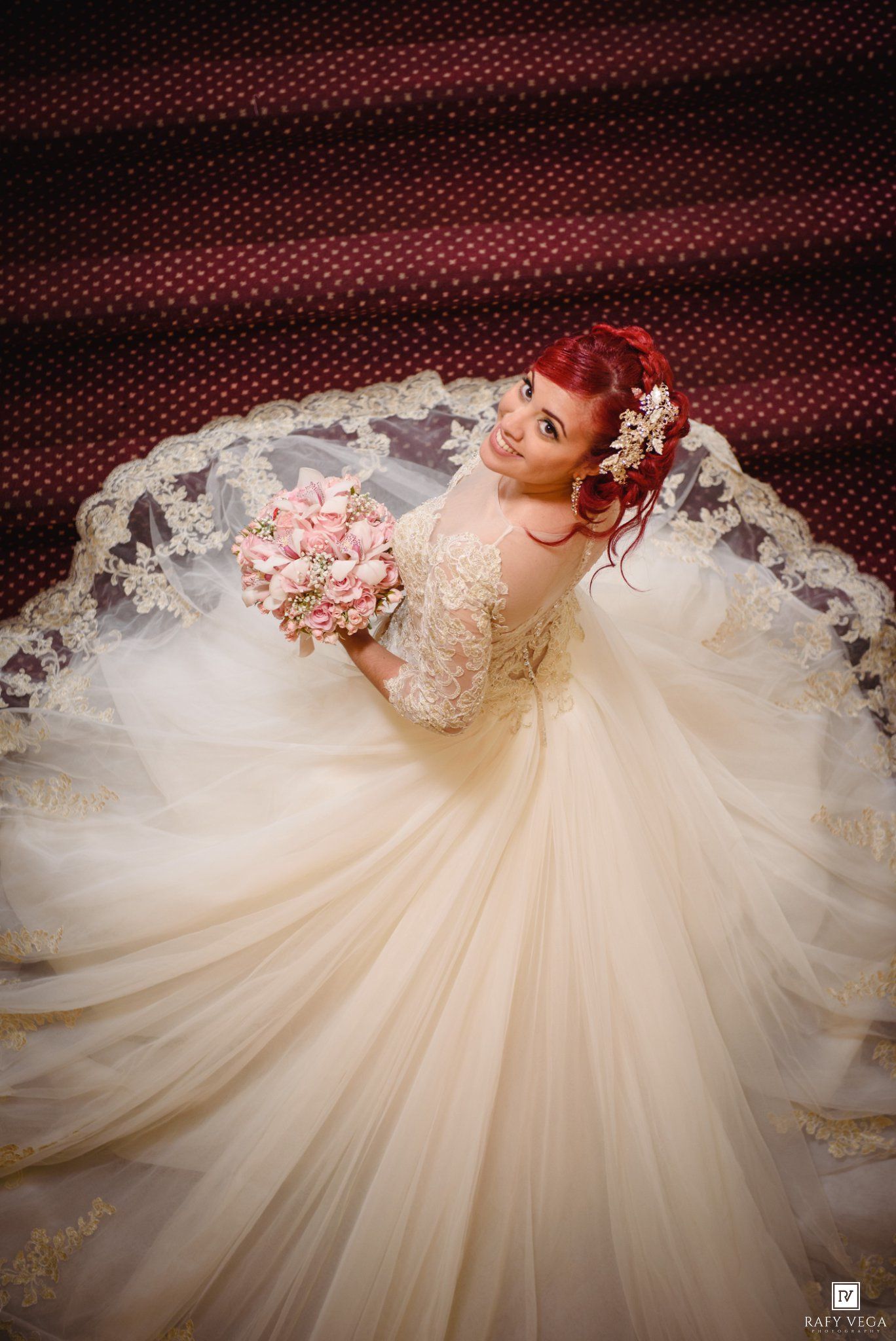 Bride in ivory lace ball gown with gold trim holds pink bouquet, red hair adorned with flowers, veil spread elegantly.