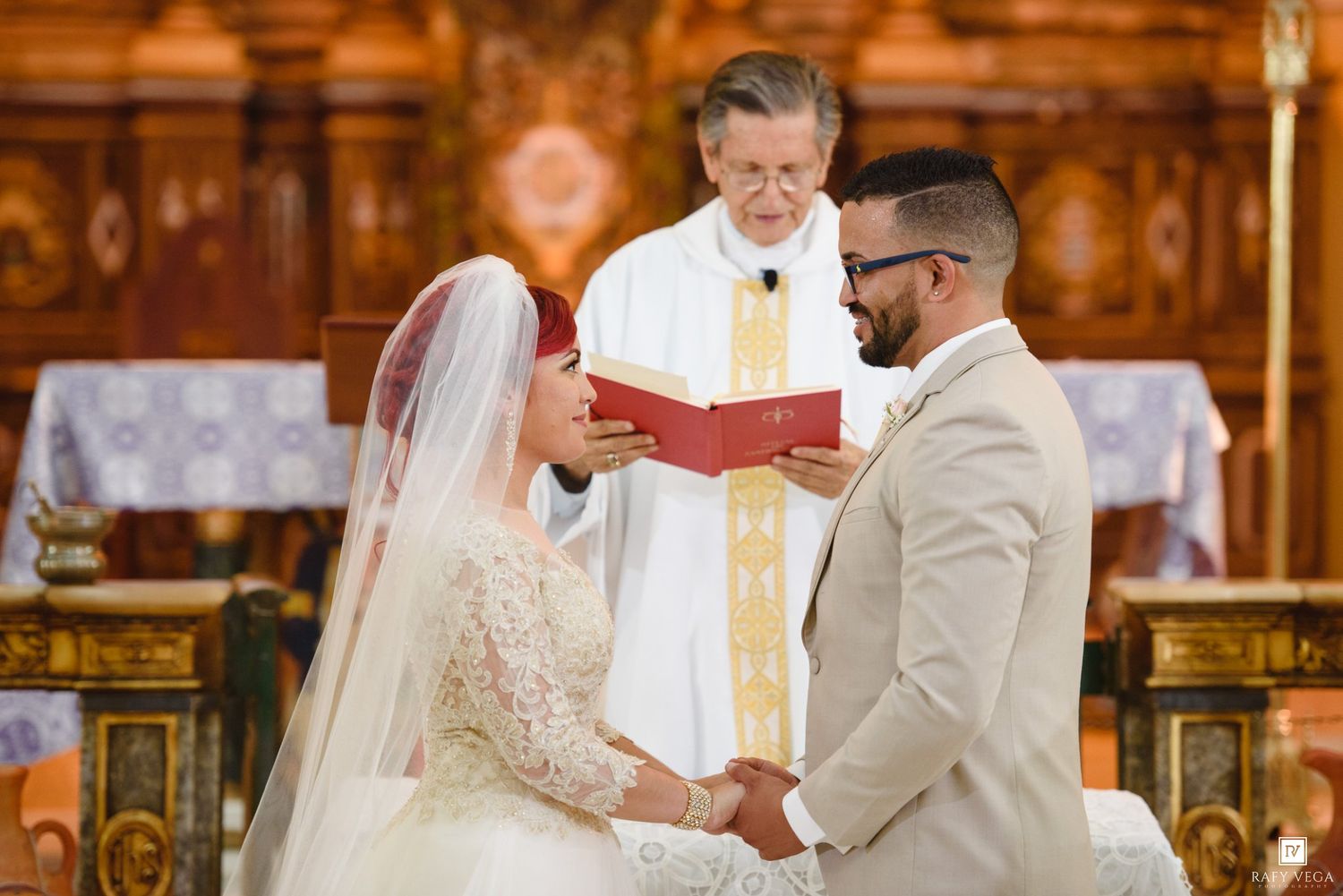 Bride and groom exchange vows at church altar with priest holding red book during wedding ceremony.