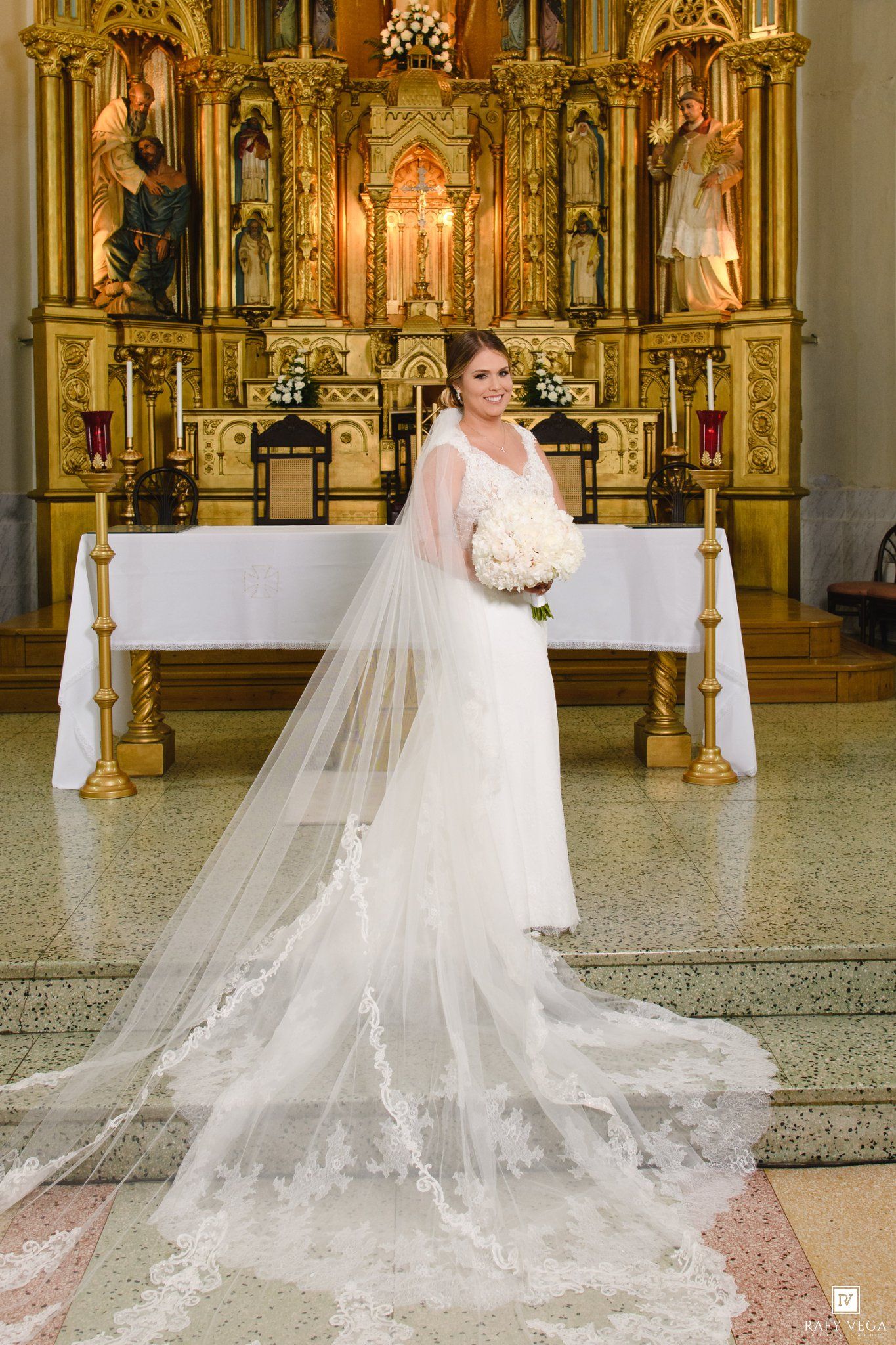 Bride in elegant lace wedding gown with long cathedral veil stands before golden altar in ornate church.