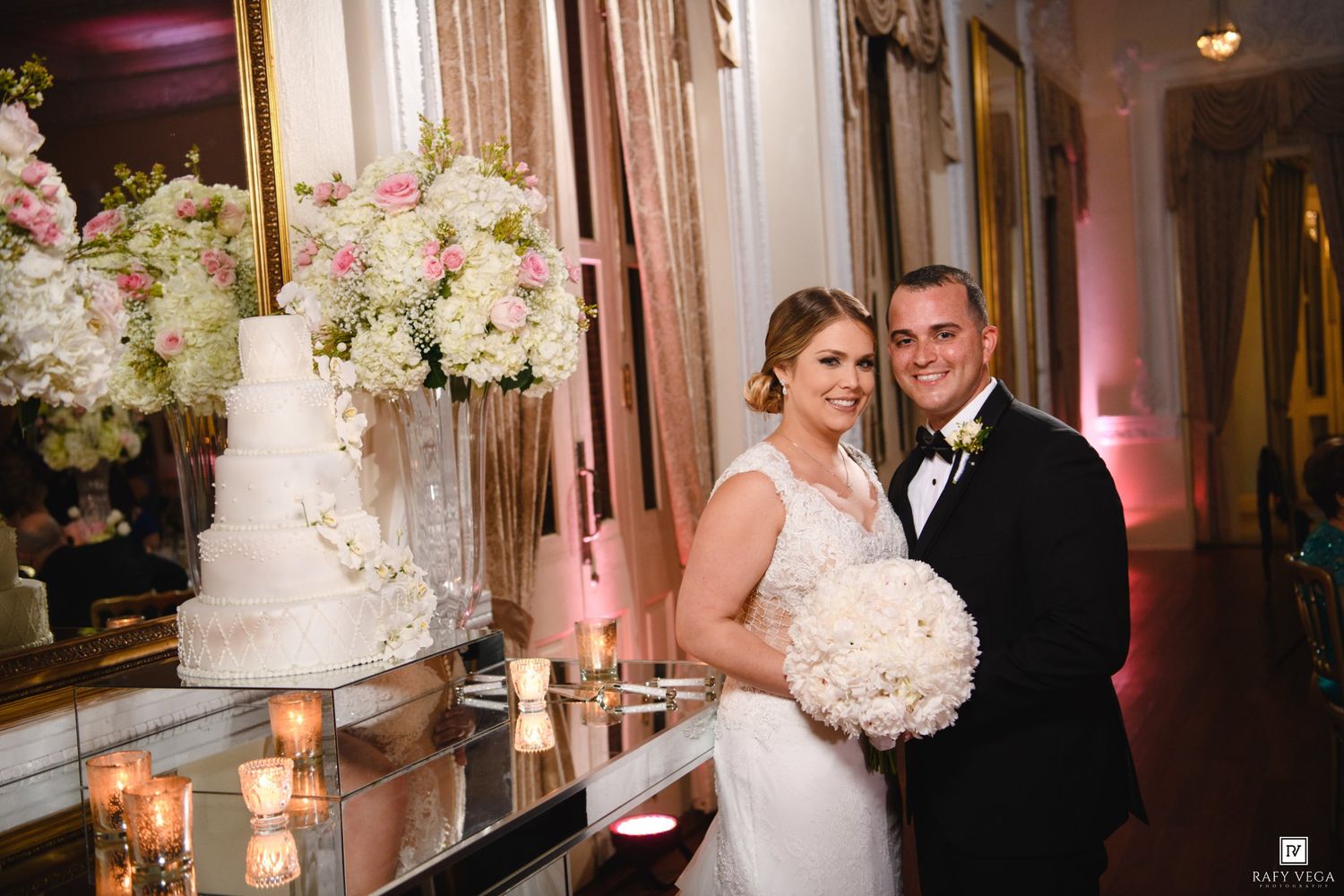 Bride and groom smiling beside a elegant white wedding cake with floral arrangements at a romantic reception venue.