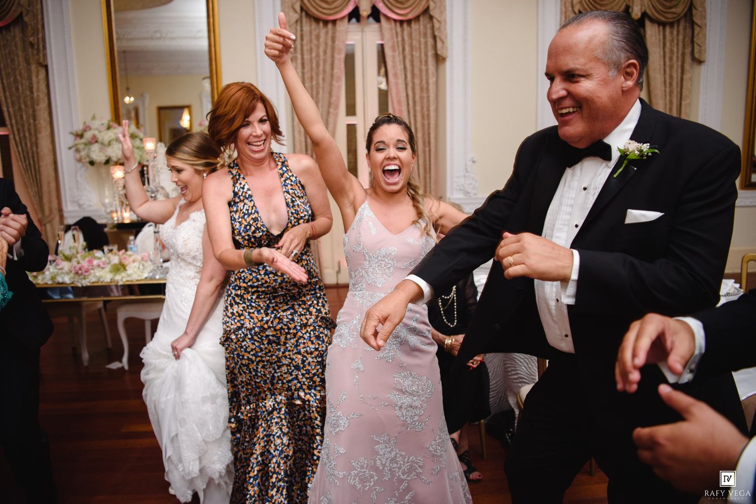 Guests dance and celebrate joyfully at a wedding reception, with the bride visible in a white gown.
