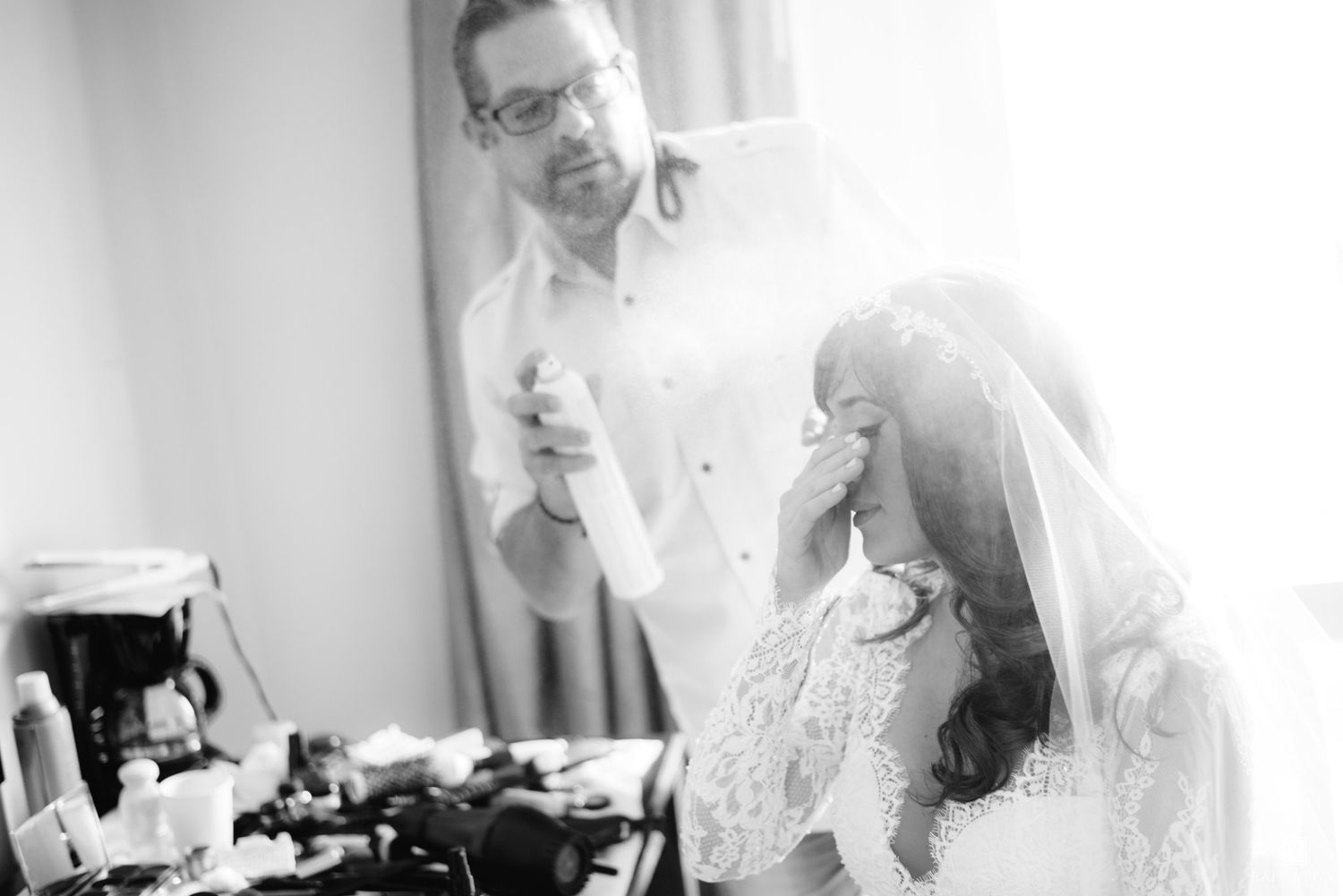 Hairstylist sprays bride's veil as she wipes tears in emotional black and white wedding preparation photo.