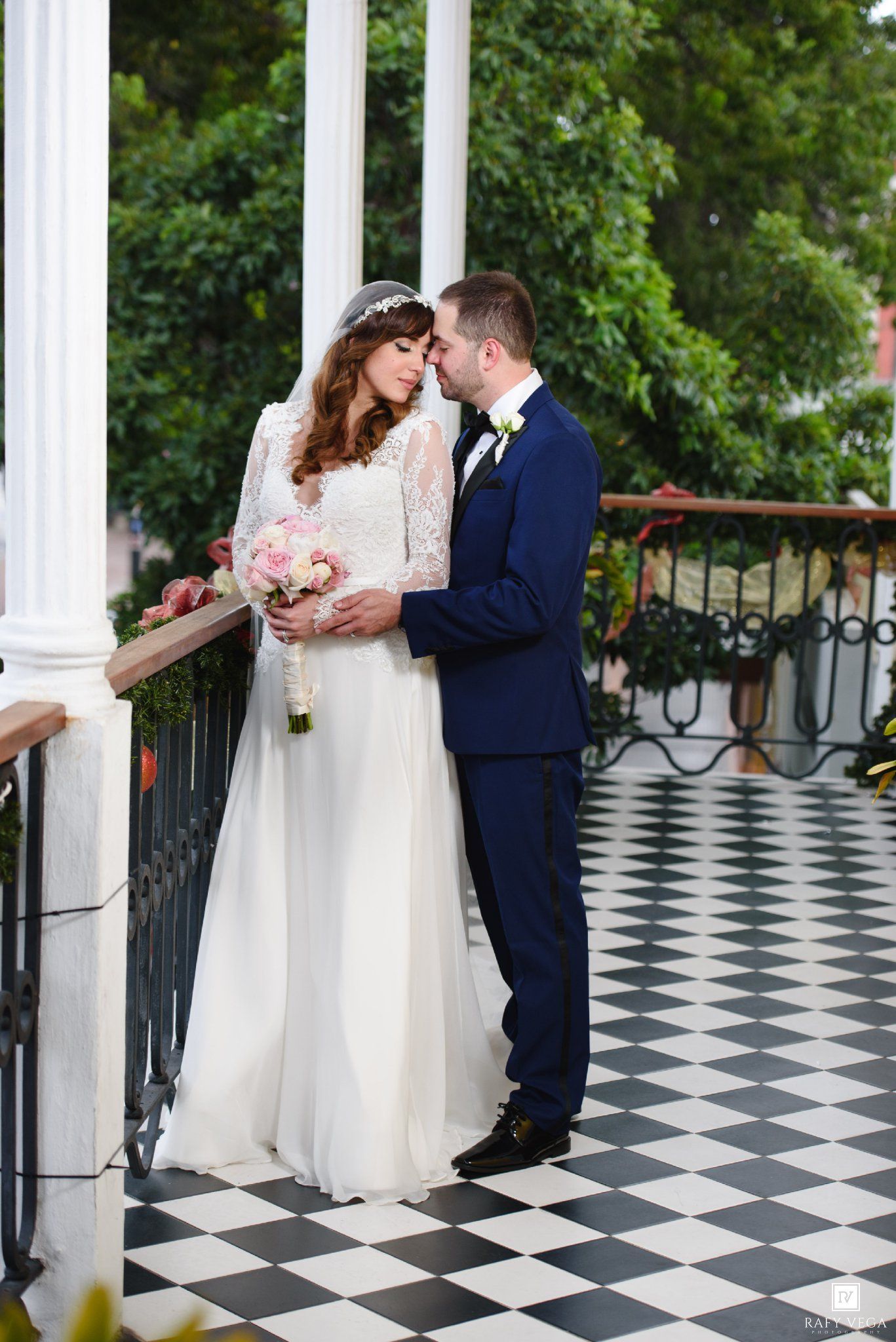 Bride and groom share a romantic moment on a checkered balcony, bride holding pink bouquet in lace wedding gown.