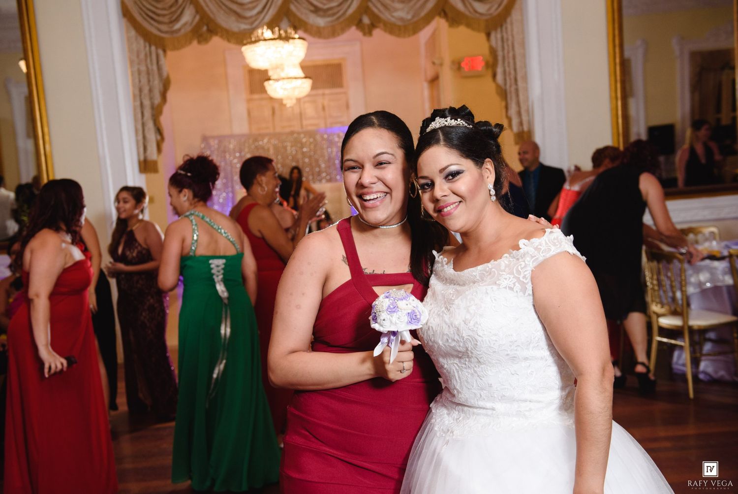 Bride in white lace gown poses with bridesmaid in red dress at elegant wedding reception venue.