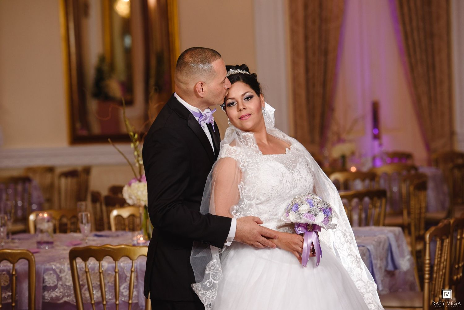 Bride in white lace gown holding purple bouquet stands with groom in black tuxedo in elegant banquet hall.