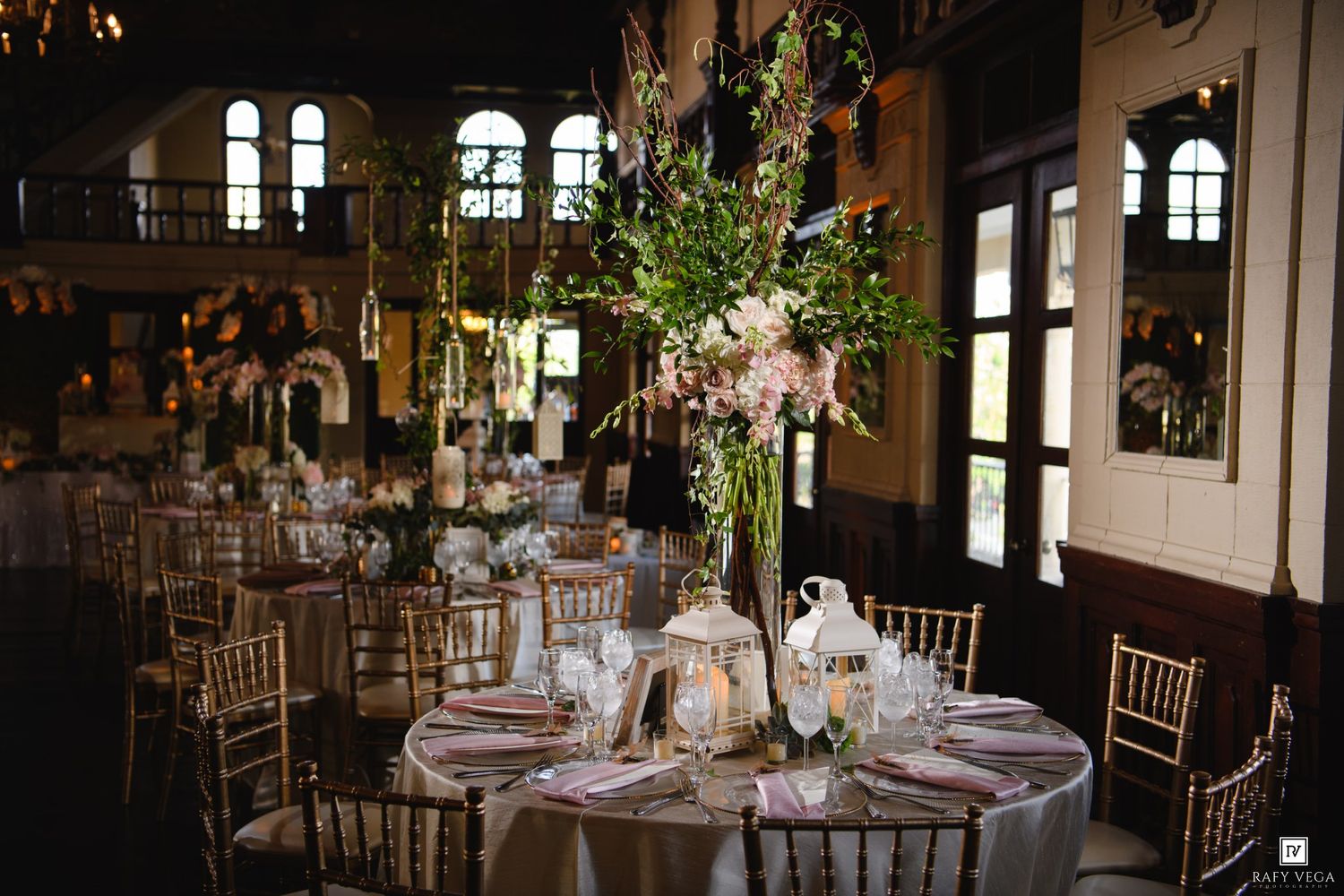 Elegant wedding reception hall with tall floral centerpieces, gold chiavari chairs, and round tables with pink linens.
