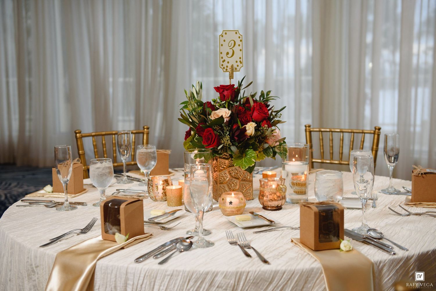 Elegant wedding reception table with red rose centerpiece, gold chiavari chairs, candles, and white linen tablecloth.
