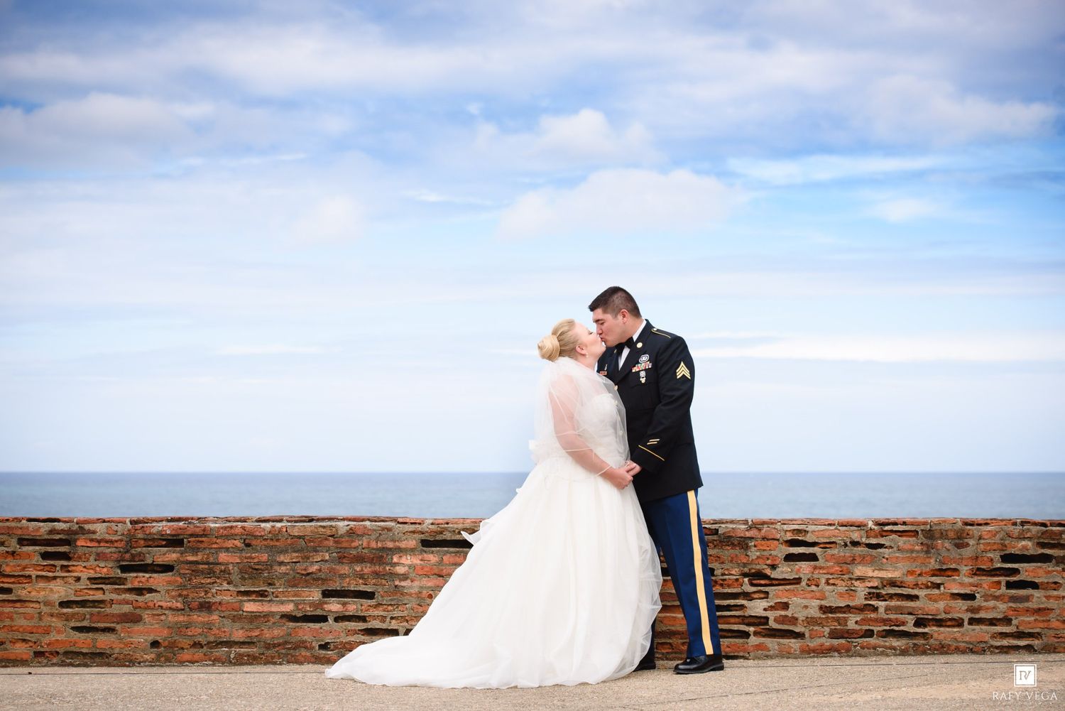 Military groom in dress uniform kisses bride in white gown by brick wall with ocean backdrop on wedding day.