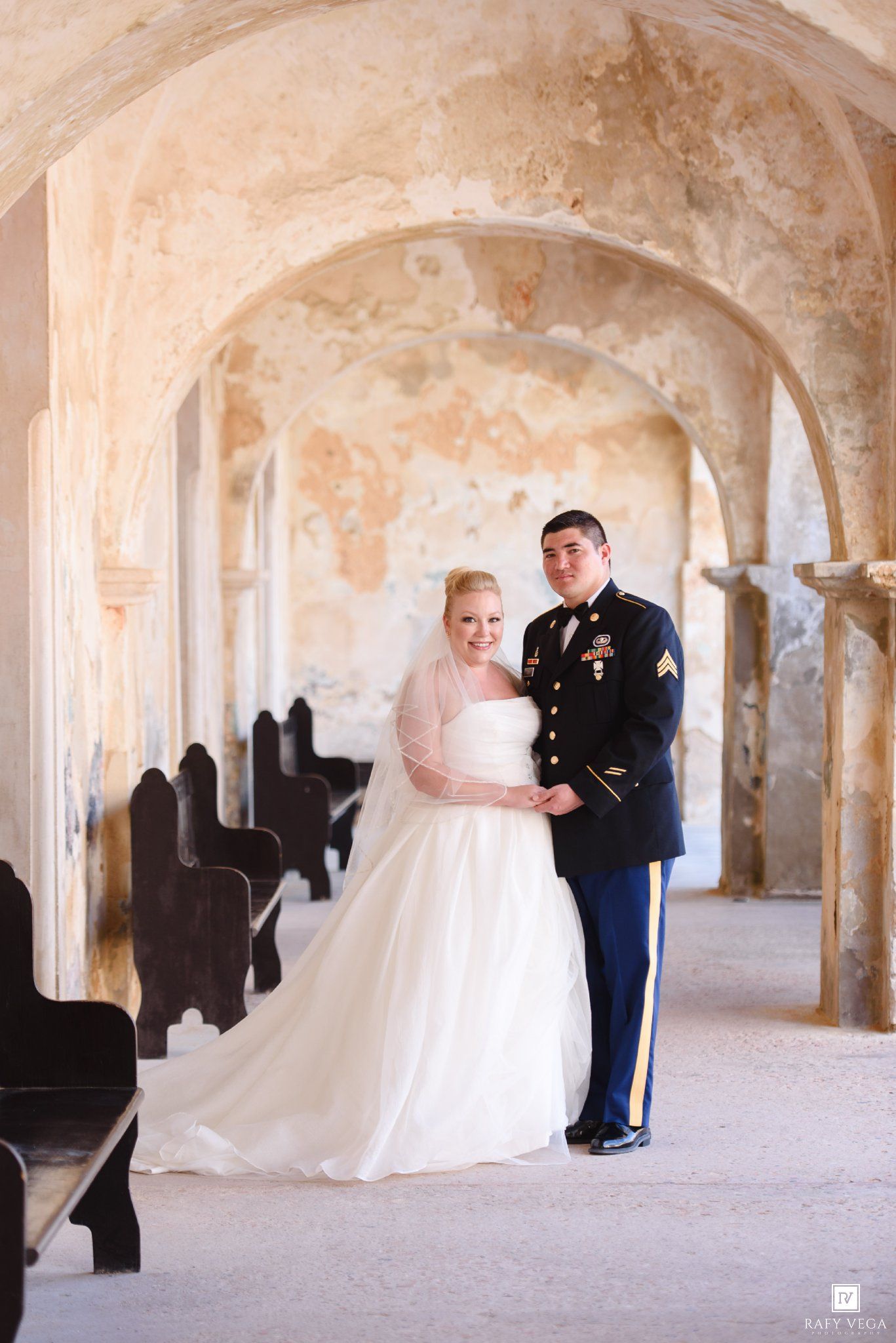 Military groom in dress uniform stands with bride in white gown under stone arches at historic wedding venue.