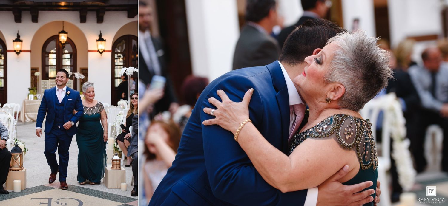 A groom in a navy suit embraces his mother warmly at an elegant wedding reception venue.