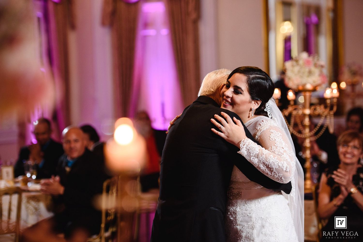 Bride in lace gown shares emotional hug with guest at elegant candlelit wedding reception with purple lighting.