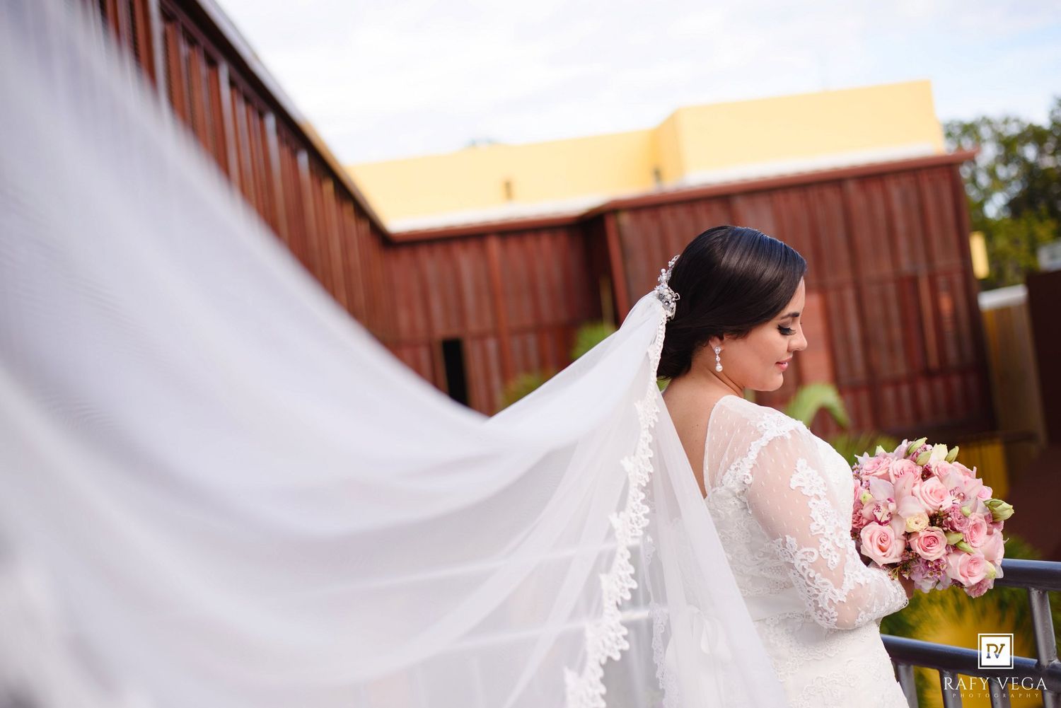 Bride in lace wedding gown with flowing veil holds pink bouquet, standing on balcony with building backdrop.