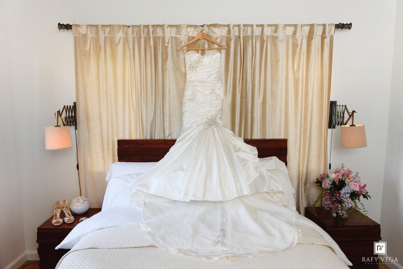 Elegant white mermaid wedding dress hanging on a hanger above a bed with floral bouquet and heels nearby.