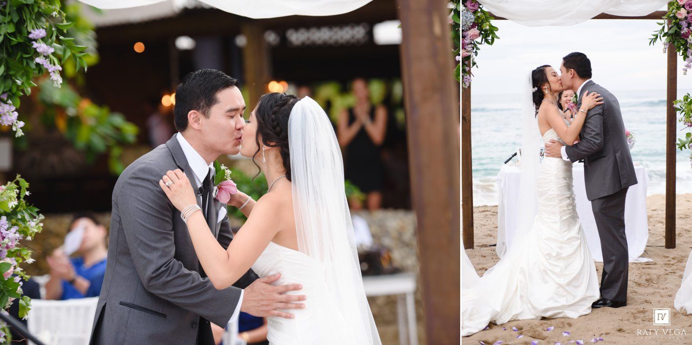 Couple shares first kiss as newlyweds at beachside wedding ceremony under floral arch.