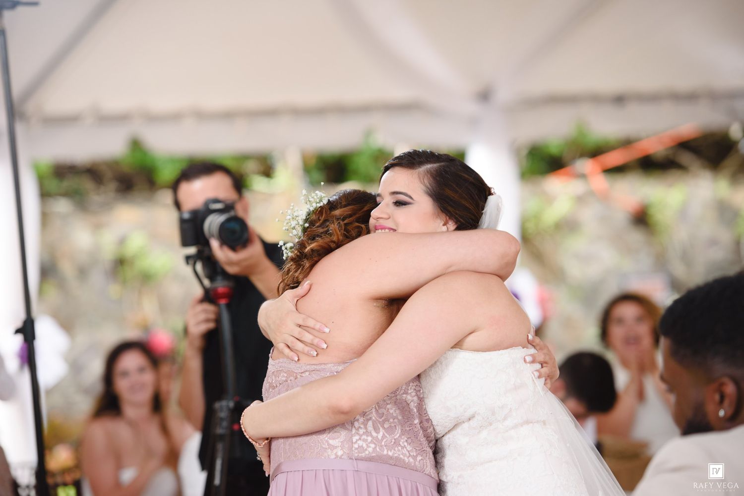 Bride and bridesmaid share an emotional hug at outdoor wedding reception under white tent.