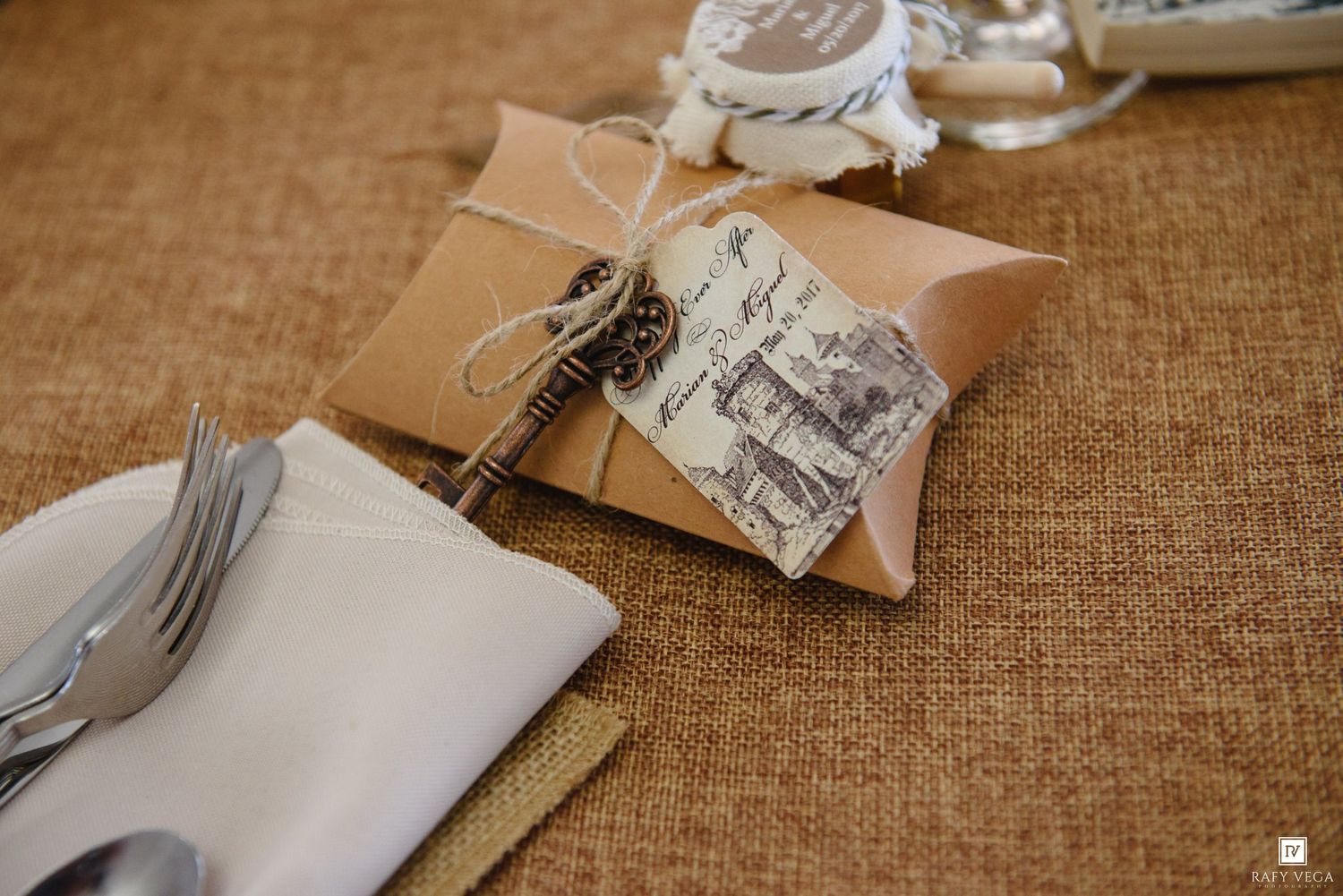 Rustic wedding favor in kraft paper pillow box with vintage key and postcard tag on burlap table runner.