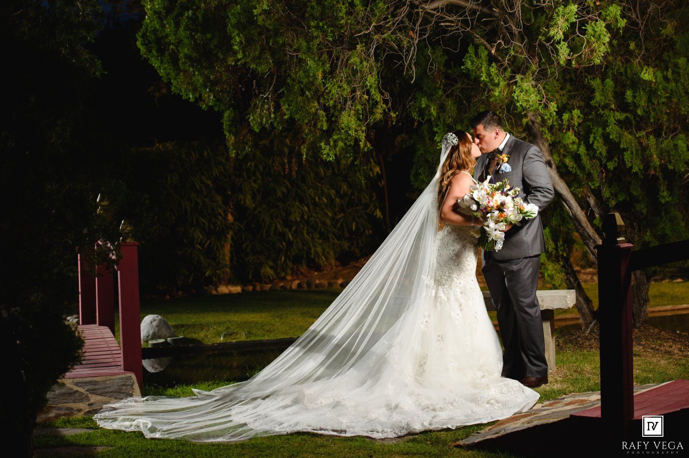 Bride and groom share a romantic kiss at night, with bride's long veil trailing on lush green garden grounds.
