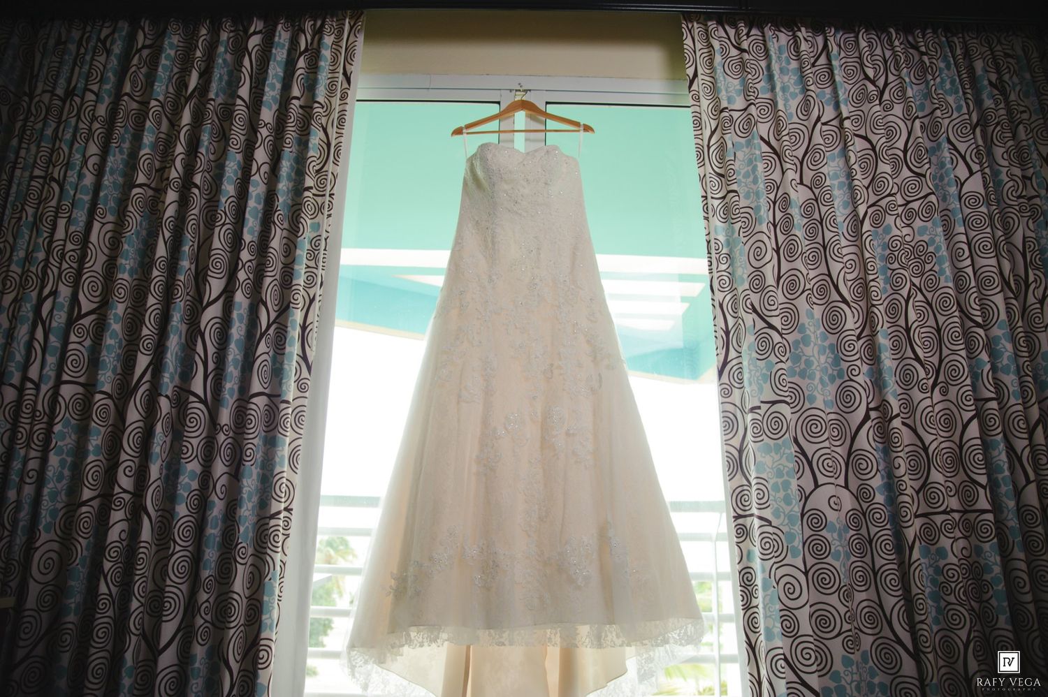 Elegant white lace wedding dress hanging on a wooden hanger between ornate blue curtains by a bright window.