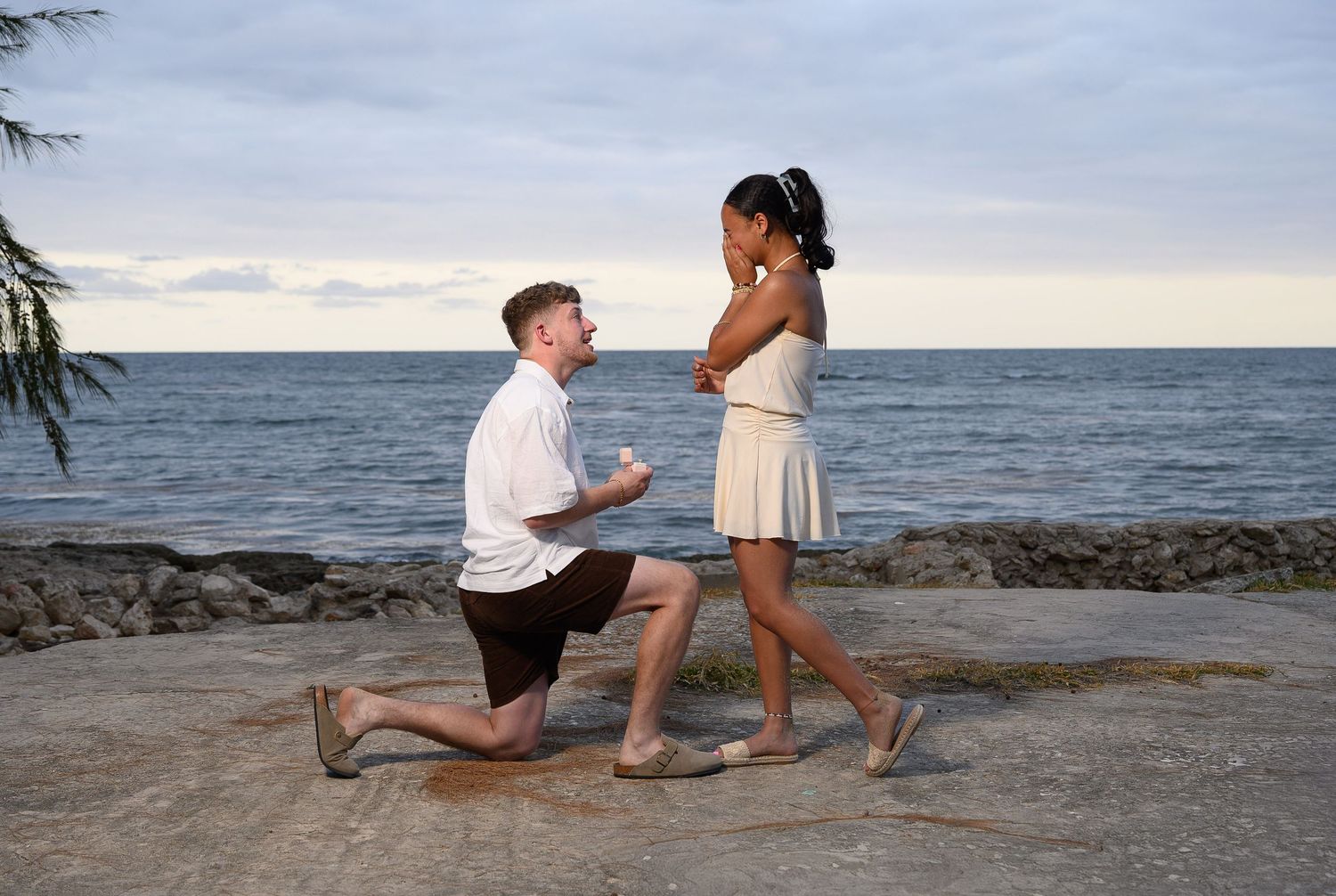 Man proposing to woman on one knee by the ocean at sunset, romantic beach marriage proposal scene.