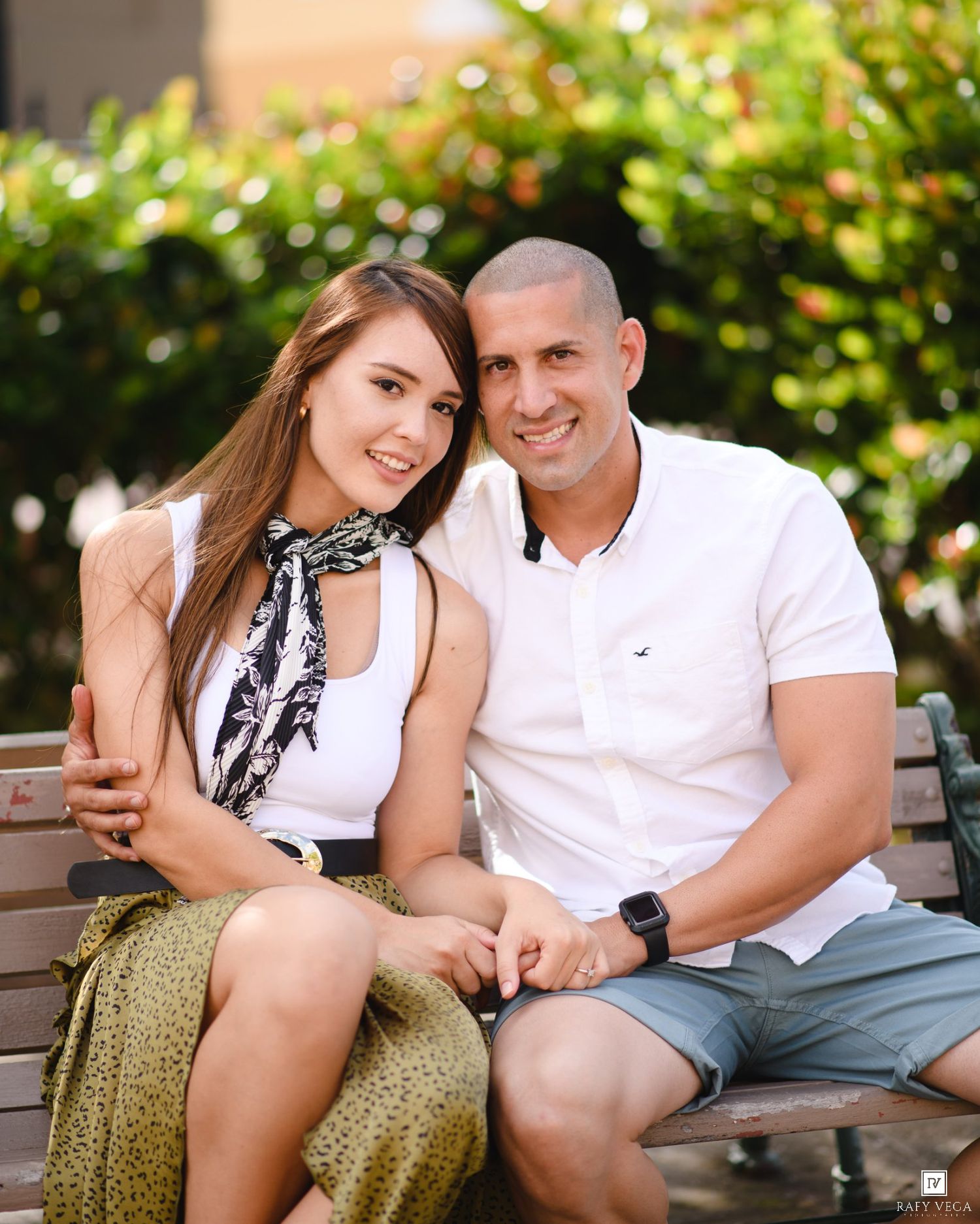 A smiling couple sits on a park bench surrounded by greenery, with the man in a white polo embracing the woman.