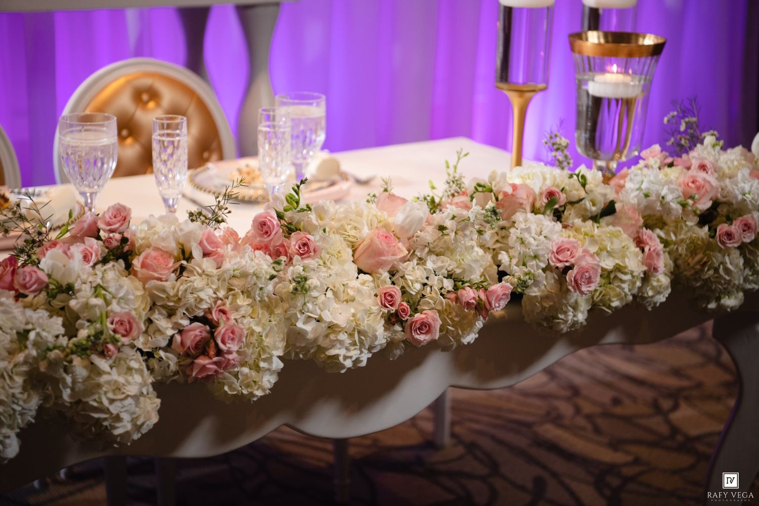 Elegant wedding head table adorned with lush pink roses and white hydrangeas floral garland with crystal glassware.