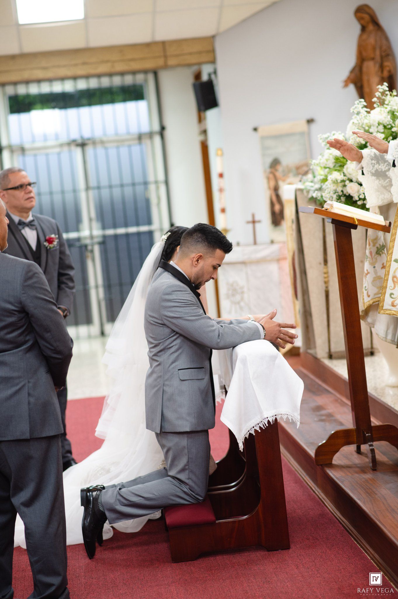 Bride and groom kneeling at church altar during Catholic wedding ceremony, groom in gray suit, bride in white gown.