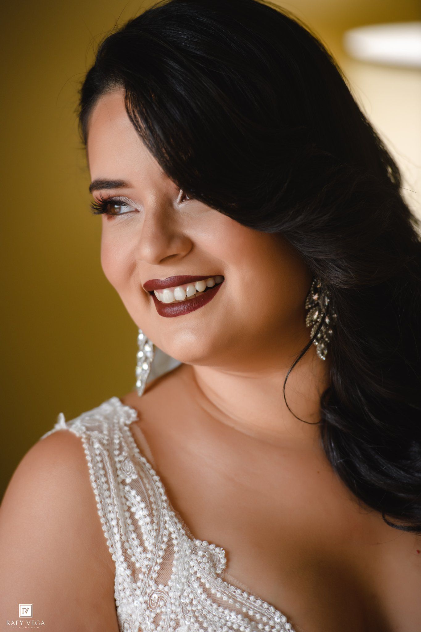 Smiling bride with dark hair, pearl earrings, and beaded white gown, wearing bold berry lipstick.