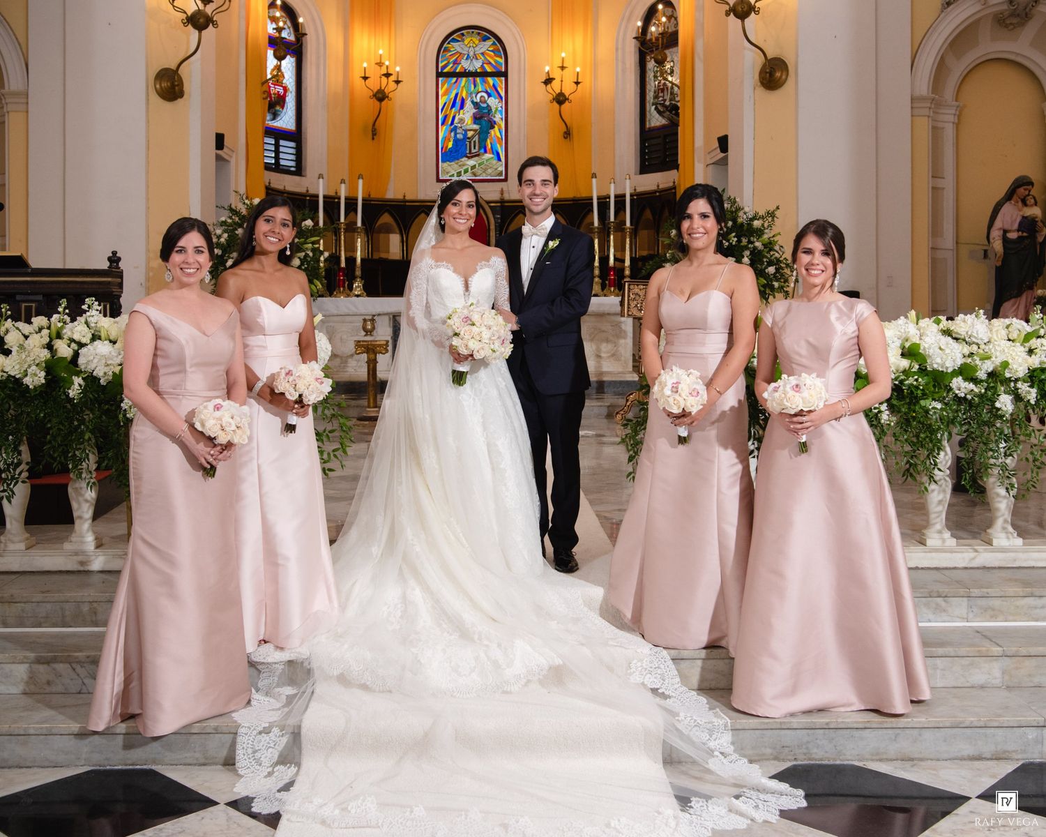 Bride and groom pose with bridesmaids in blush pink gowns holding bouquets inside an elegant church.